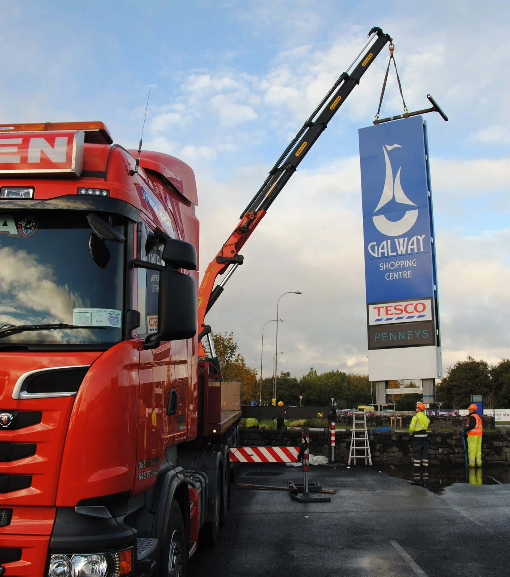 A red crane truck lifting a large sign for Galway Shopping Centre, with workers in safety vests and helmets nearby.