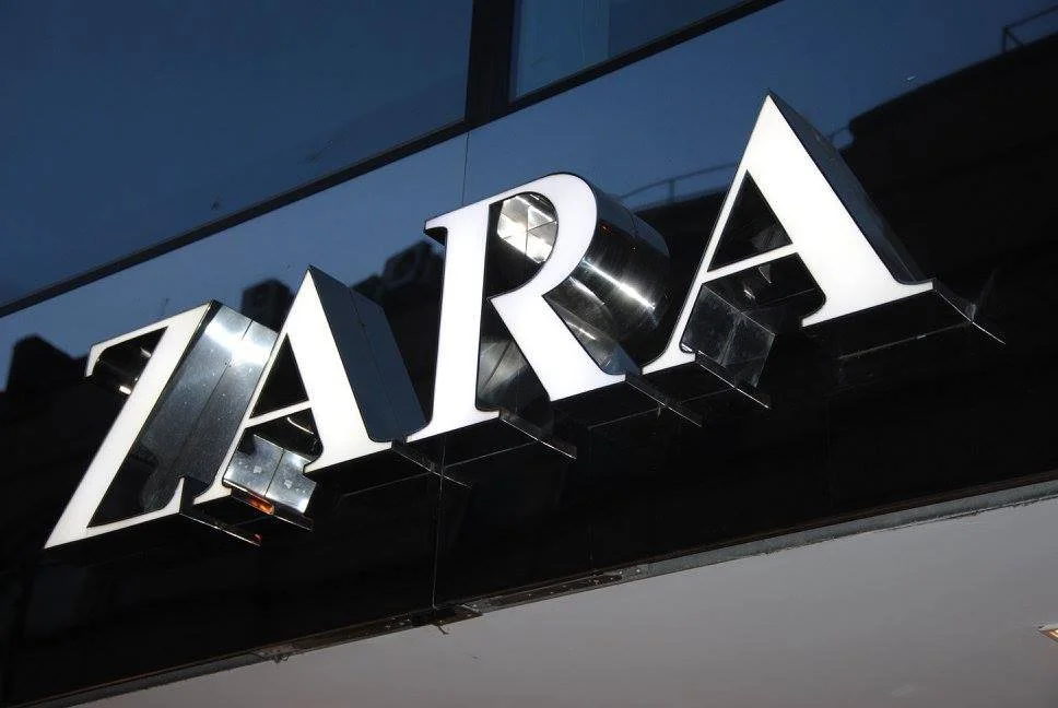 Close-up of Zara store sign with illuminated white letters on a black background at dusk.