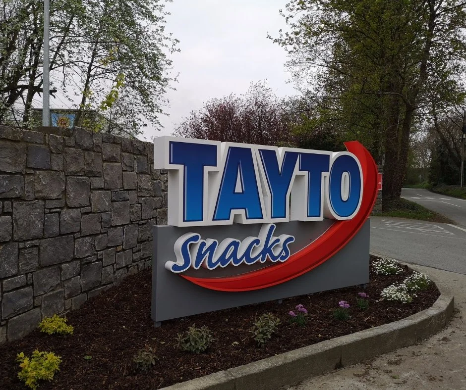 A sign for TAYTO Snacks with blue and white text, a red swoosh, and a gray background, situated next to a stone wall and a curved sidewalk, with trees and a road in the background.