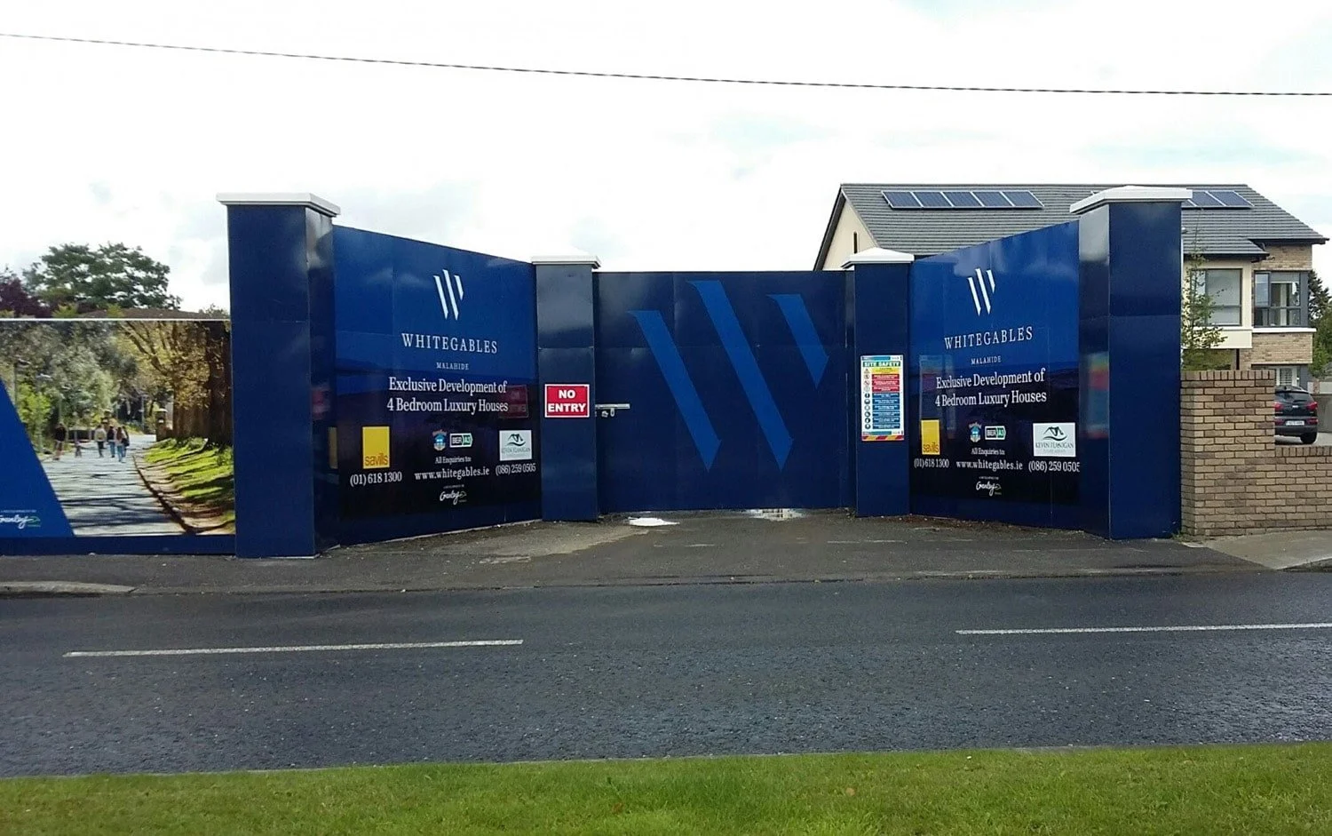 Blue construction fence with signs advertising Whitegables luxury houses, a 'no entry' sign, and a residential neighborhood with houses and parking lot in the background.