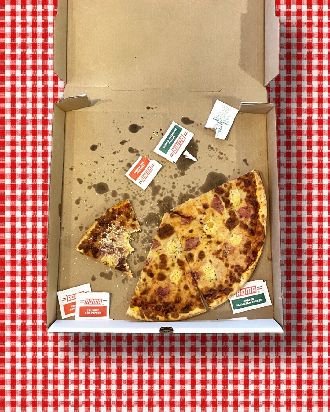 Half-eaten pizza in a box on a red and white checkered tablecloth, with sauce and cheese splatters and pizza toppings like pineapple and ham, along with small condiment packets and some spilled sauce.