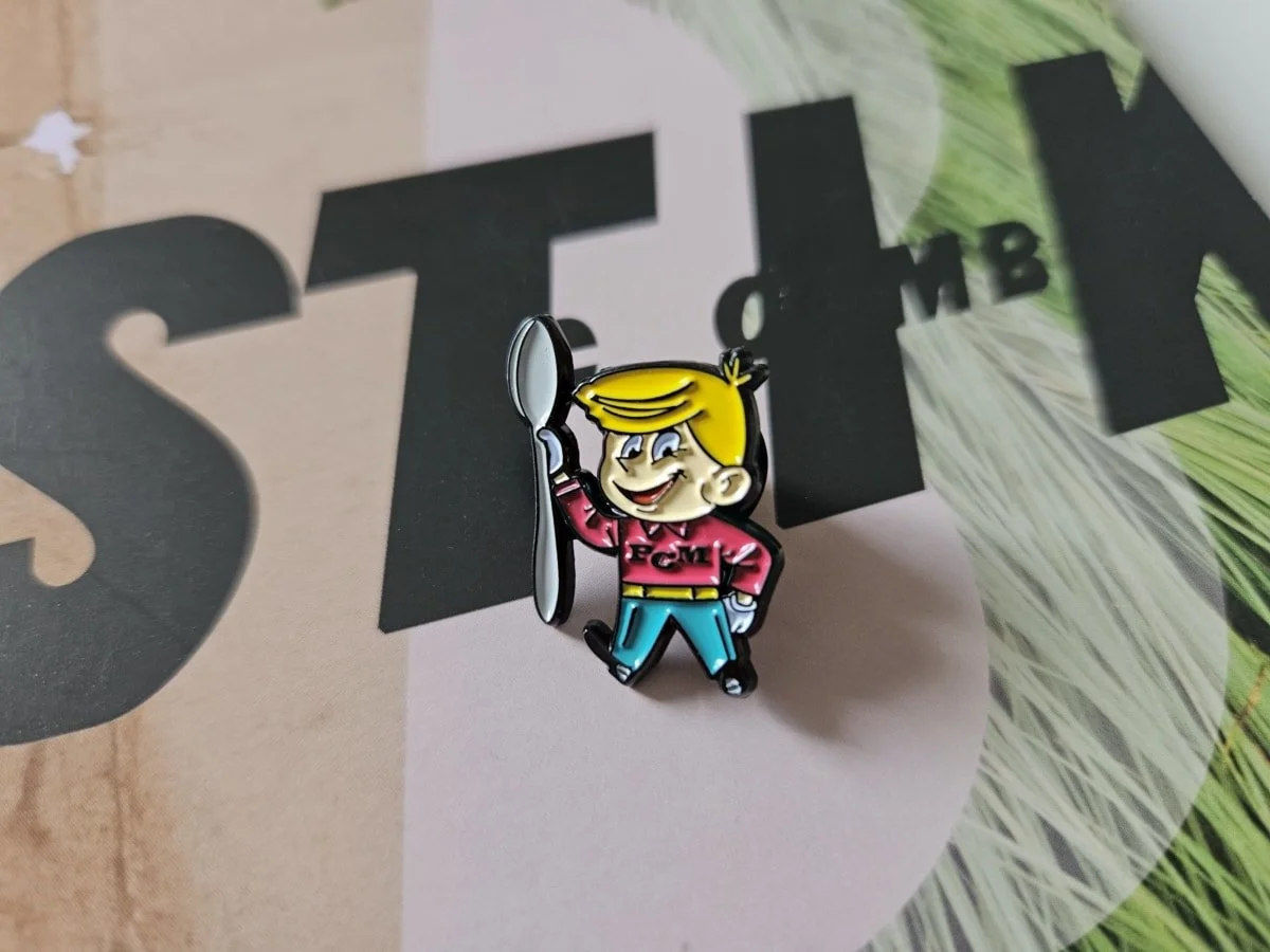 A cartoon pin of a smiling boy with blonde hair, wearing a red shirt with 'FCM' on it, blue pants, and holding a spoon, placed on a surface with mostly black and white text and some green and pink elements.