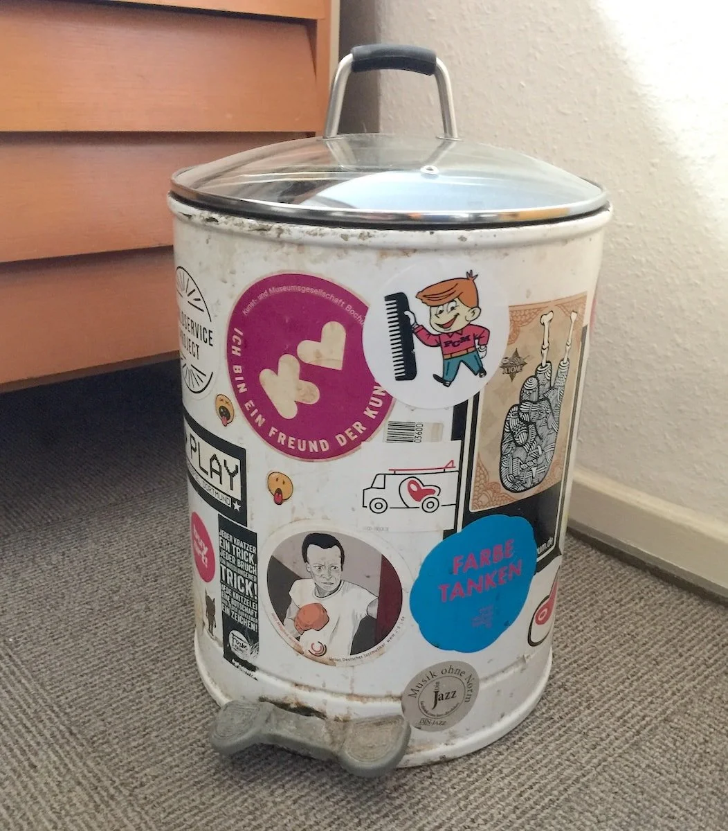 A white metal trash can covered with various colorful stickers, placed on a gray carpeted floor near a wall and a wooden surface.