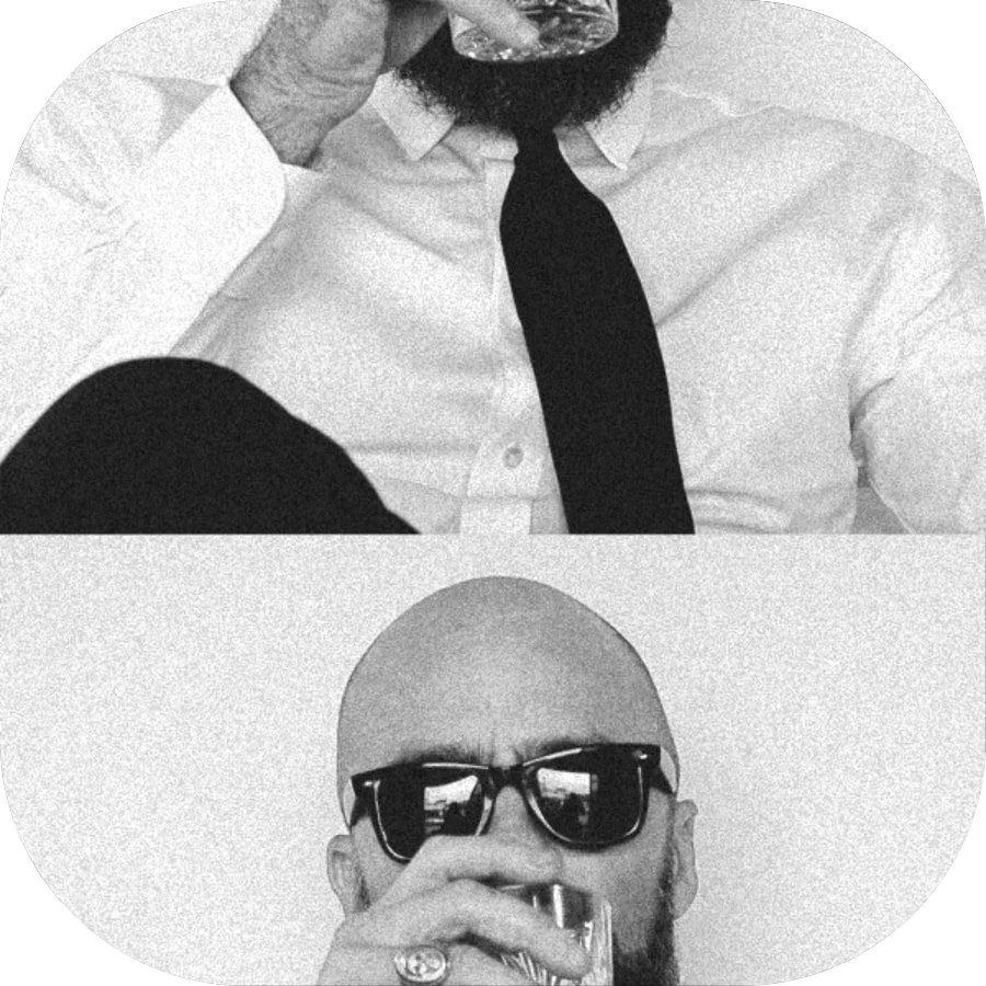 Two men drinking whiskey, one with a beard wearing a white shirt and black tie, the other with sunglasses and no hair, both holding glasses up to their lips.