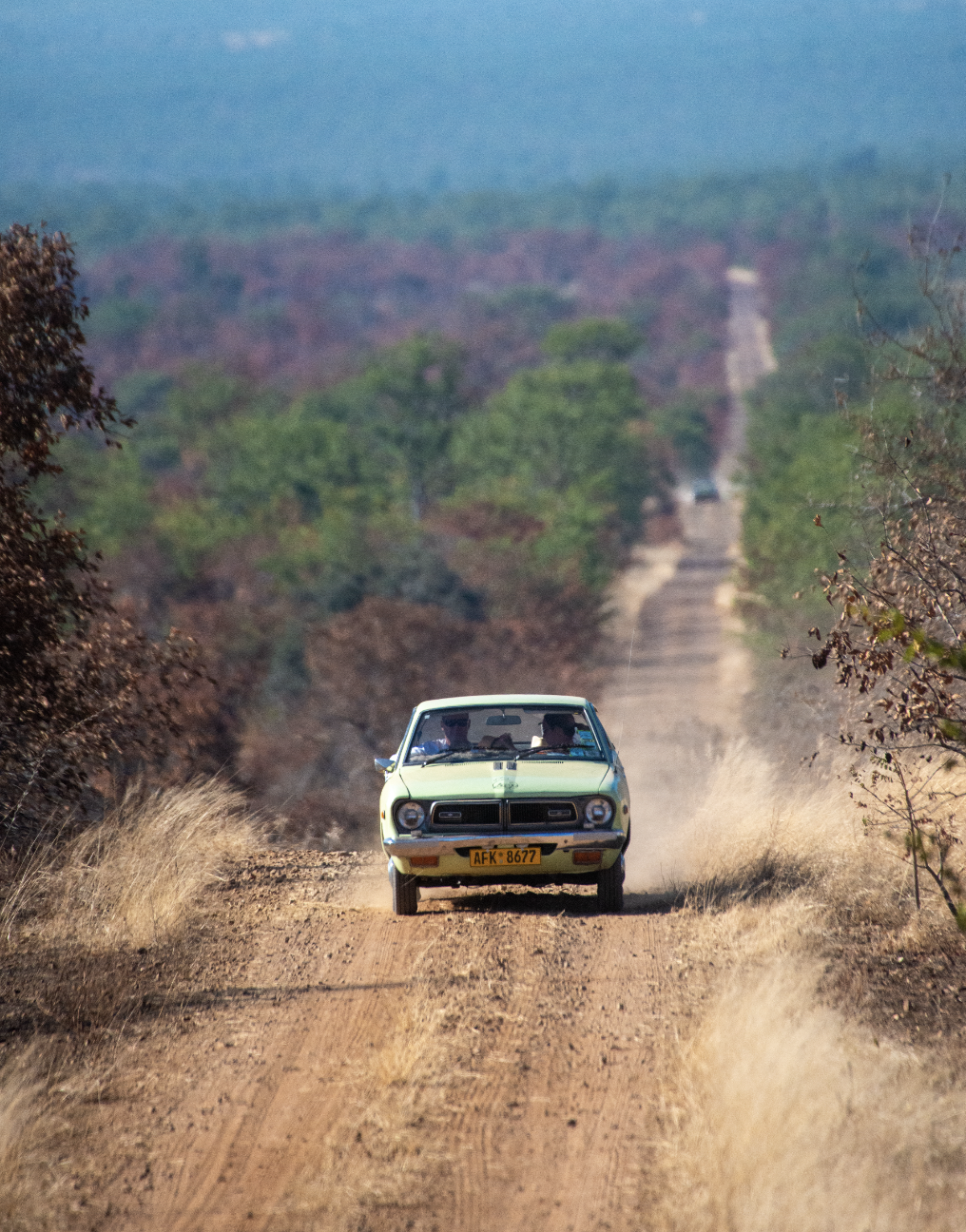 A vintage yellow car driving on a dirt road through a rural landscape with trees and a distant hill.