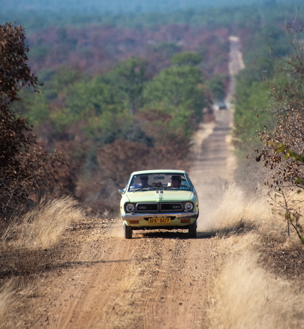 A vintage yellow car driving on a dirt road through a rural landscape with trees and distant hills.