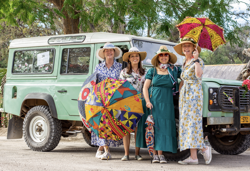 Four women standing next to a vintage green off-road vehicle outdoors, wearing summery dresses and wide-brimmed hats, holding colorful umbrellas, smiling at the camera.