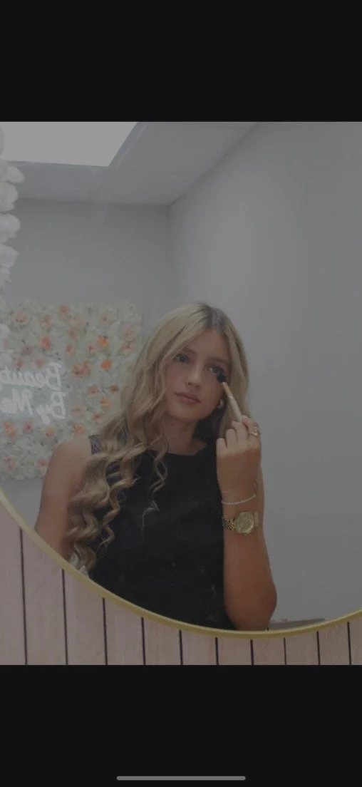 Young woman with blond hair and a black top standing in front of a wall of pink and white roses and blossoms.