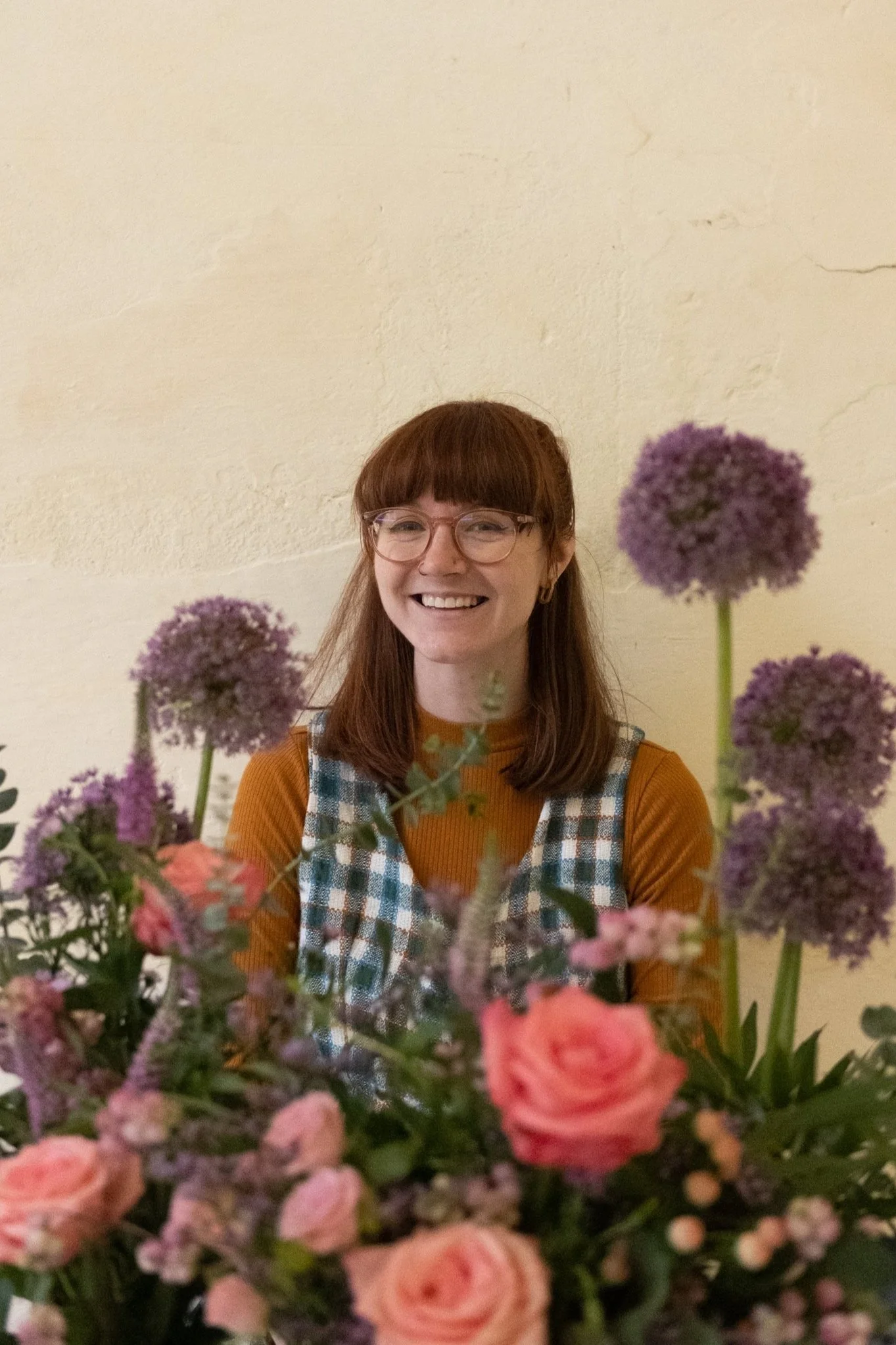 A woman with glasses and shoulder-length brown hair smiling behind a bouquet of pink and purple flowers against a plain beige wall.