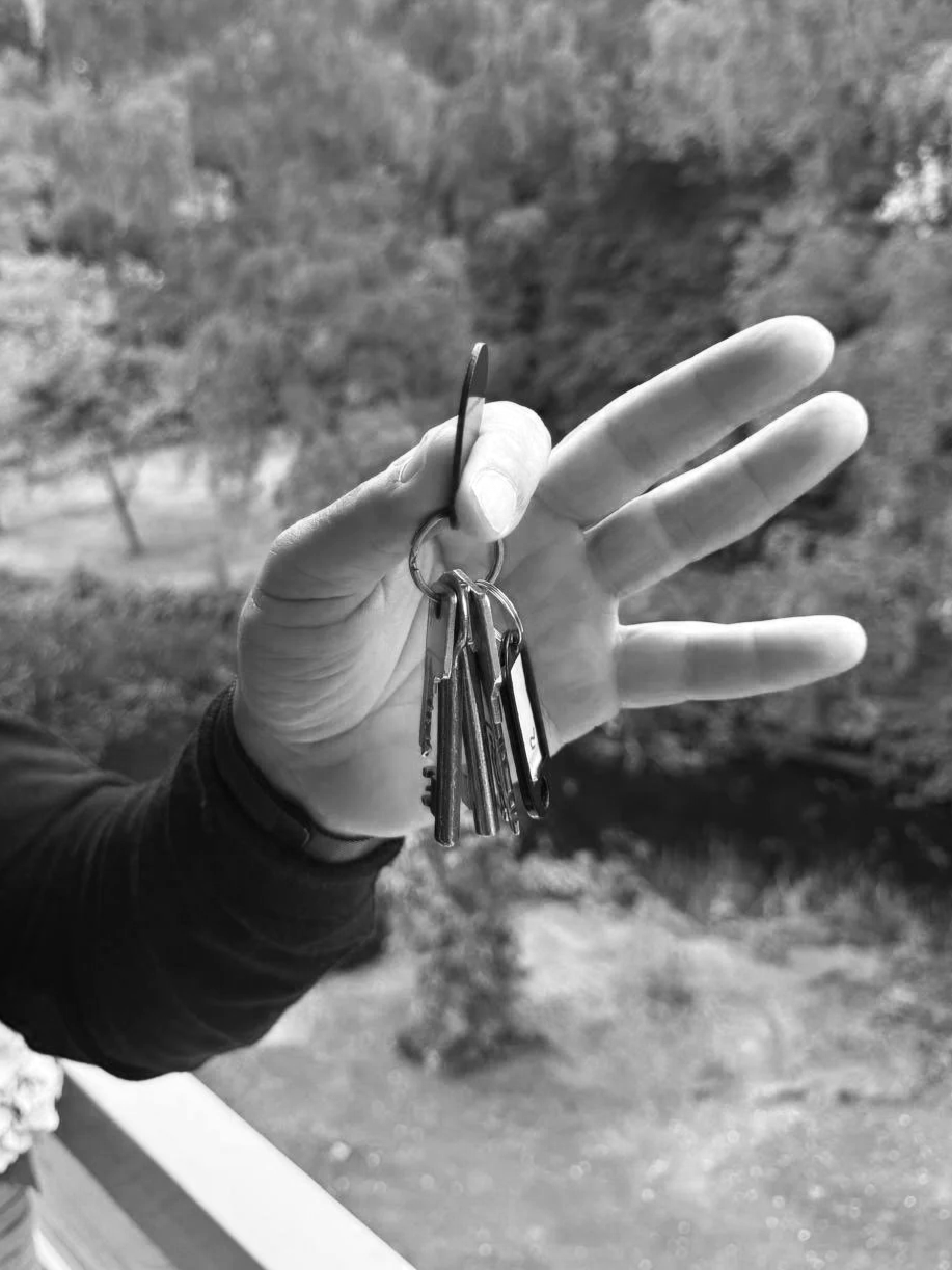 A person holding a keyring with several keys and a small screwdriver outdoors, blurred trees in the background.