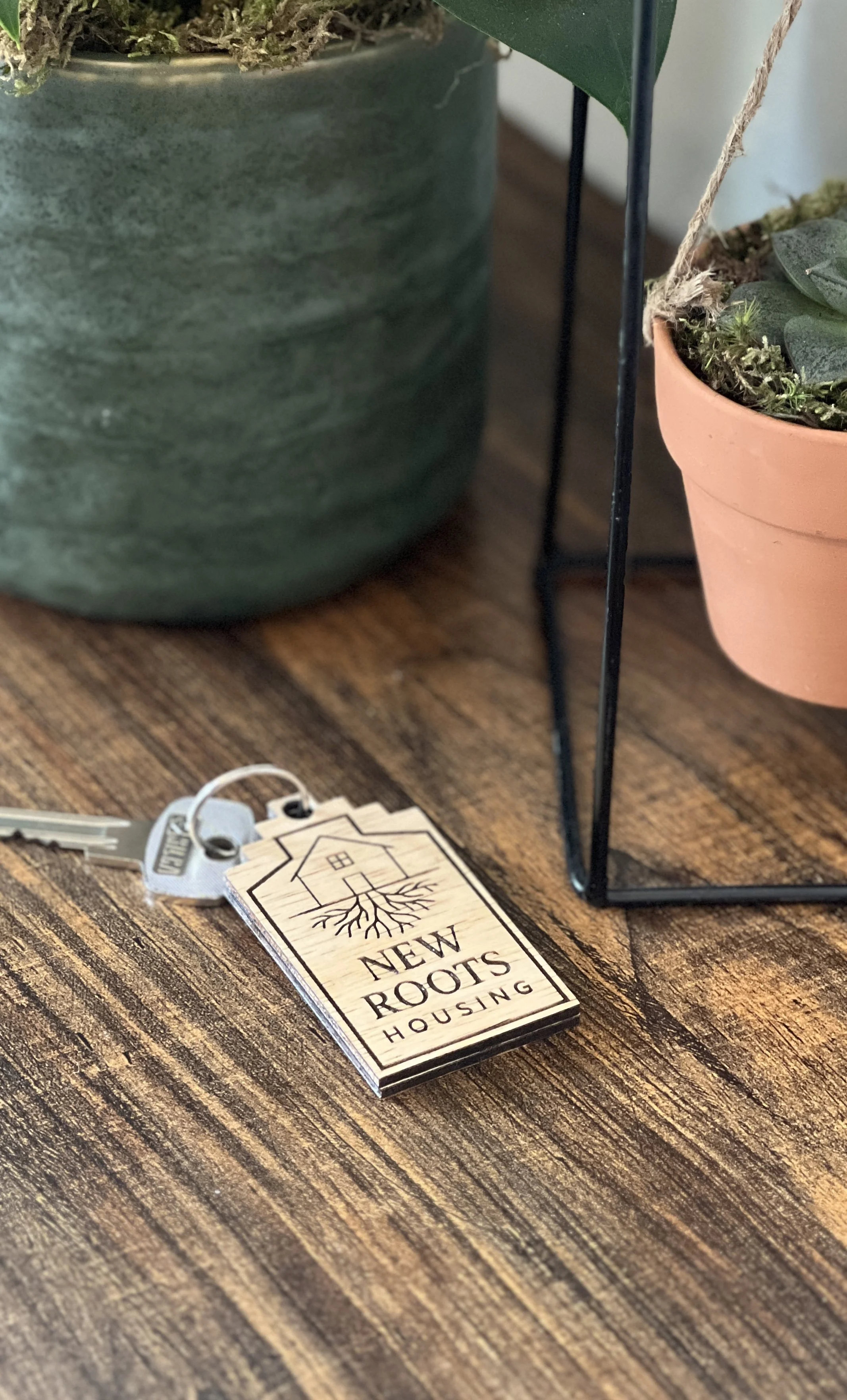 A wooden house-shaped keychain with the words 'New Roots Housing' and a tree with roots design, attached to a key, placed on a wooden surface near potted plants.