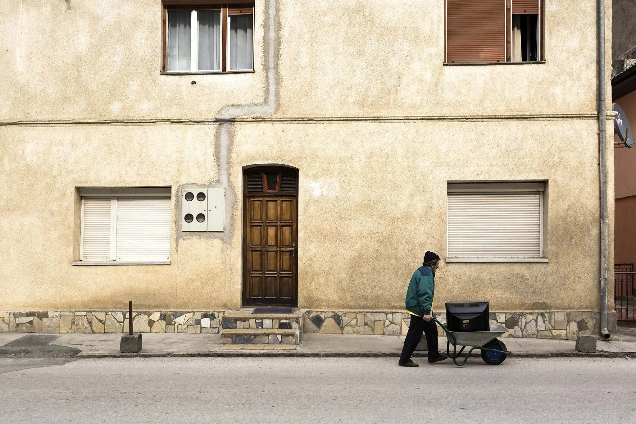 A man pushing a wheelbarrow with a large black object on a city street in front of a beige building with closed windows and a wooden door.