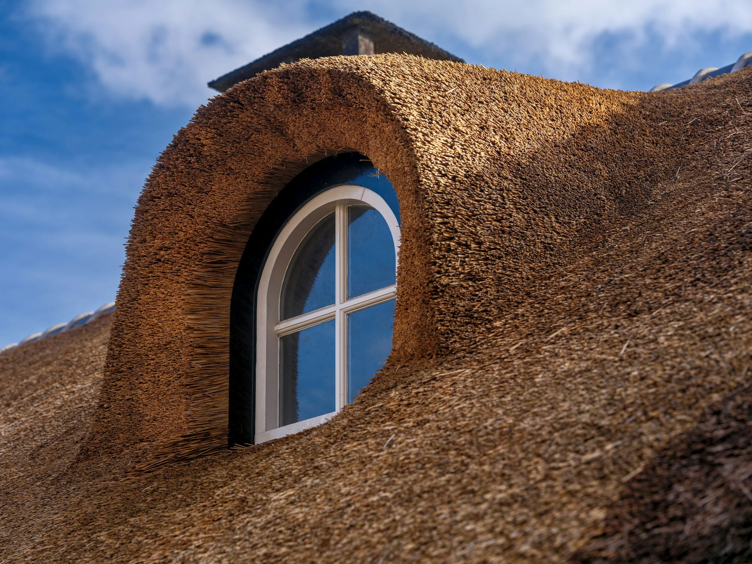 A close-up of a house with a thatched roof and a small arched window, reflecting the sky and clouds.