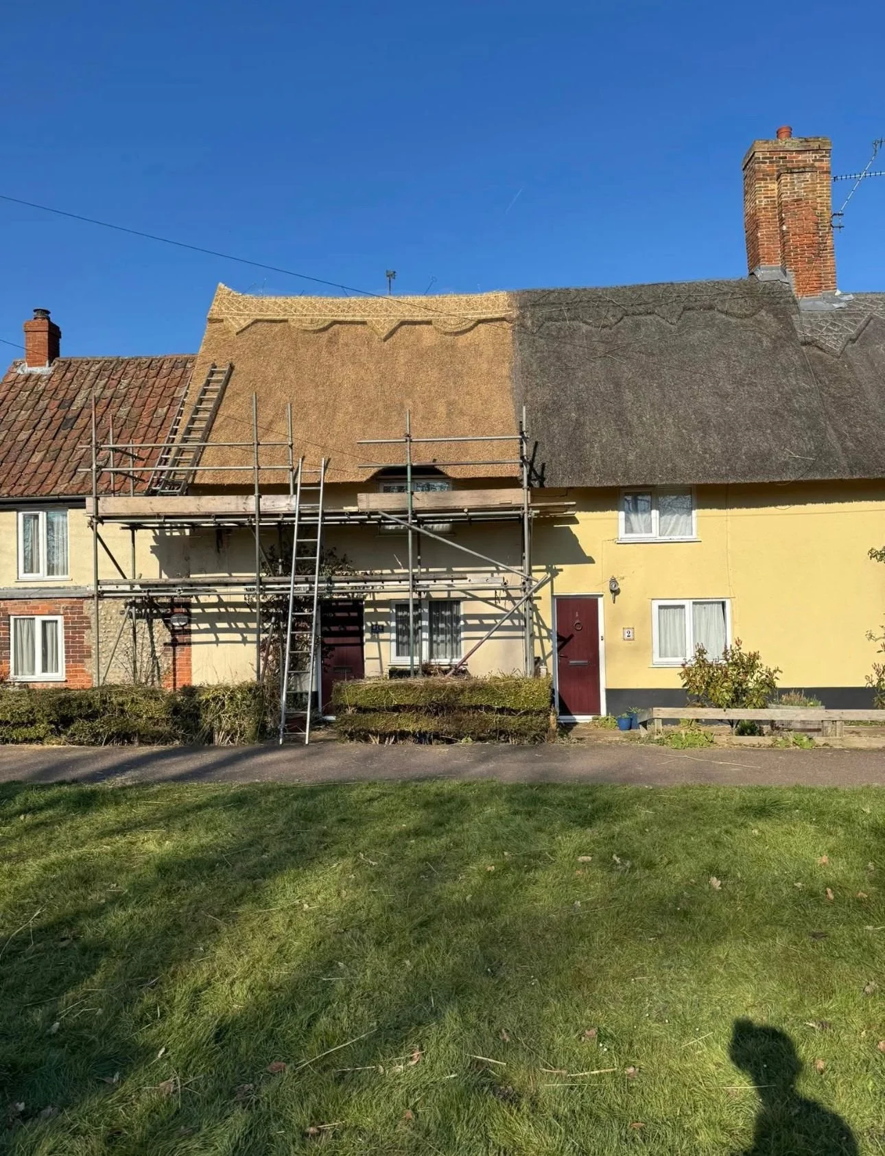 A house with a thatched roof under renovation, scaffolding set up in front, and a grassy yard with bushes and a shadow of a person taking the photo.