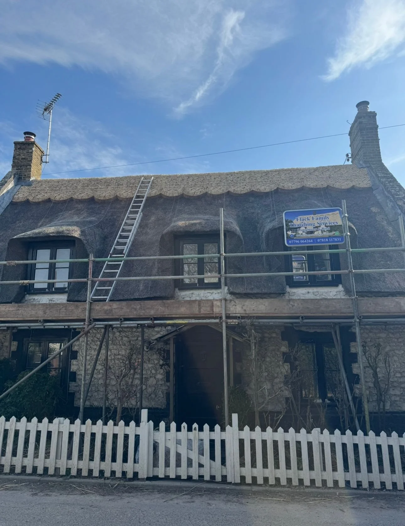House with thatched roof under renovation, scaffolding around it, ladder leaning against the roof, and a sign for Thatching Services.
