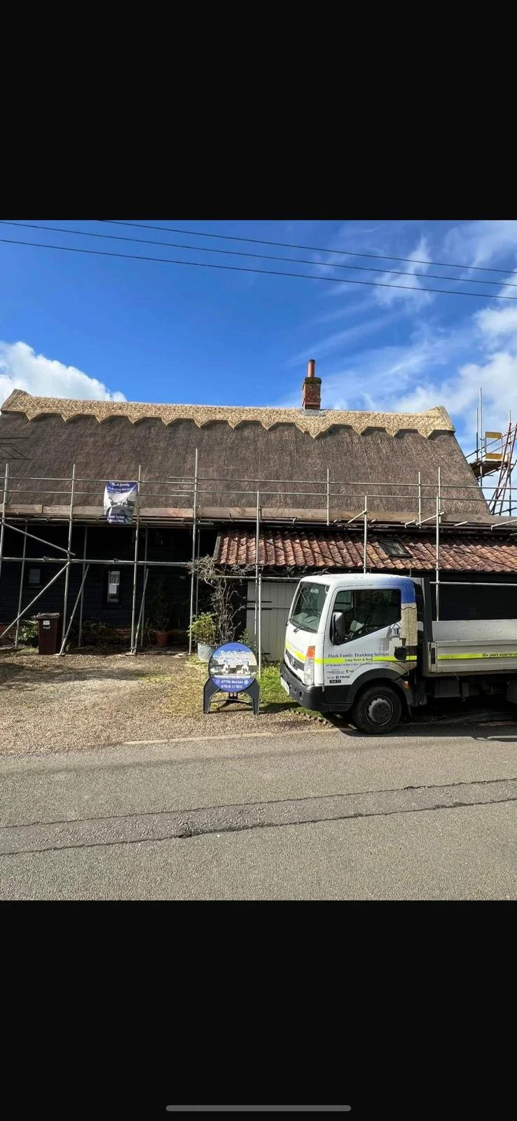 A house under renovation with scaffolding on the front, a thatched roof, and a construction vehicle parked in front. The sky is blue with some clouds.