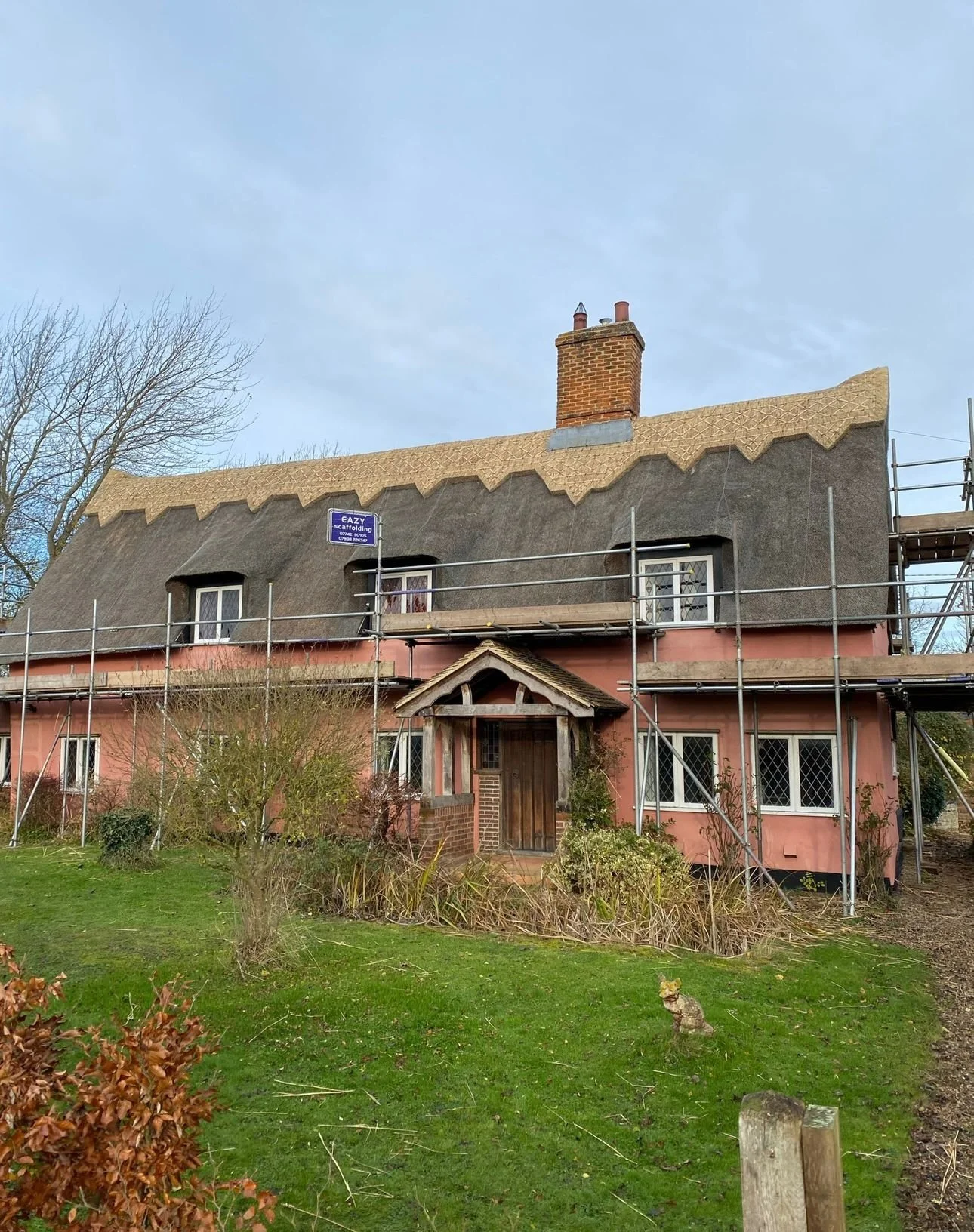A house under renovations with scaffolding, featuring a pink exterior, thatched roof, and chimney, surrounded by a grassy yard and shrubs.