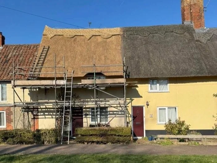 House with scaffolding and roofing work in progress, one side of the roof is rethatched while the other side has old shingles, in a residential neighborhood with a lawn and a blue sky.