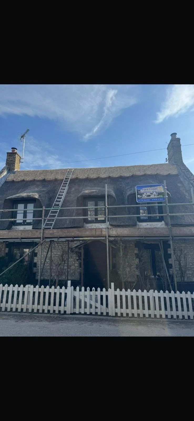 Old house under renovation with scaffolding, ladder, and a white picket fence in front. The house has a thatched roof and stone exterior, with two windows visible on the upper level. There is a sign on the scaffolding indicating construction or renovation work.