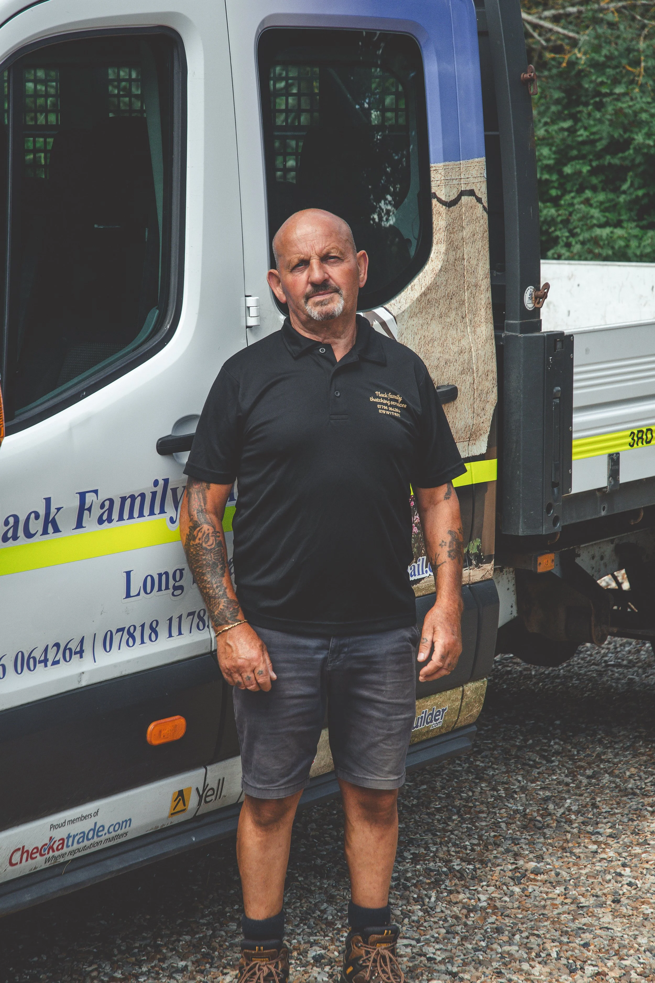 A man with tattoos on his arms standing in front of a service truck with signage for a family business, wearing a black polo shirt and shorts.