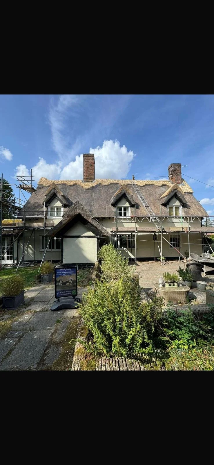 A house undergoing roof renovation with scaffolding and safety ladders in the front yard, surrounded by garden plants and outdoor furniture, under a partly cloudy sky.