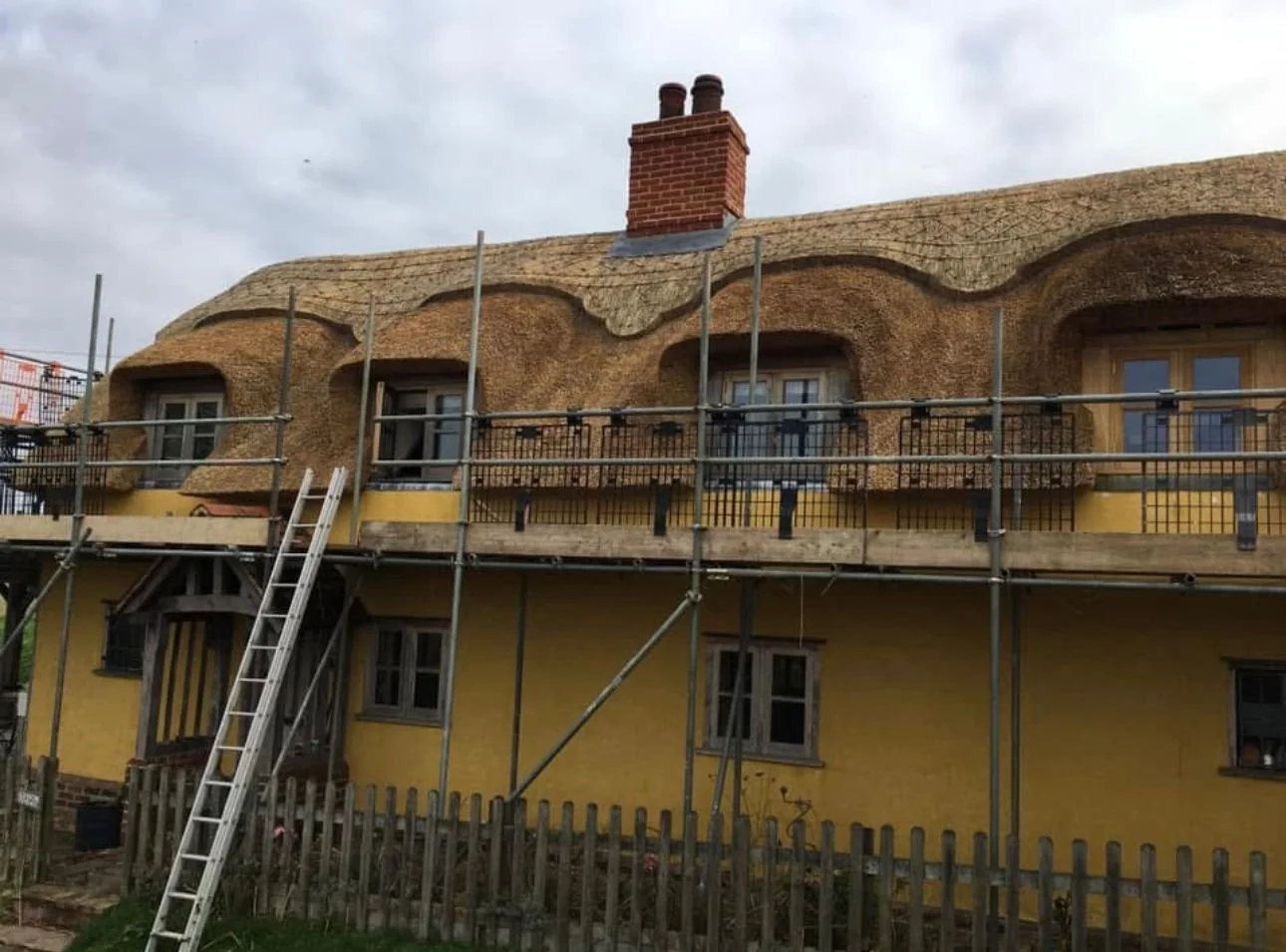 A house under construction with a thatched roof, yellow walls, and scaffolding surrounding it.