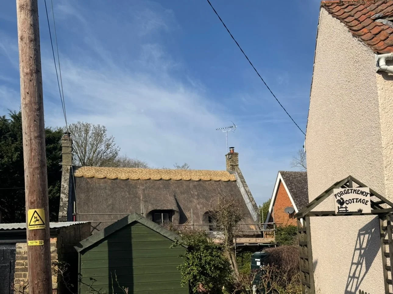 A residential area with a thatched roof cottage, a green garden shed, and a sign for Forgetmenot Cottage, under a blue sky with wispy clouds.