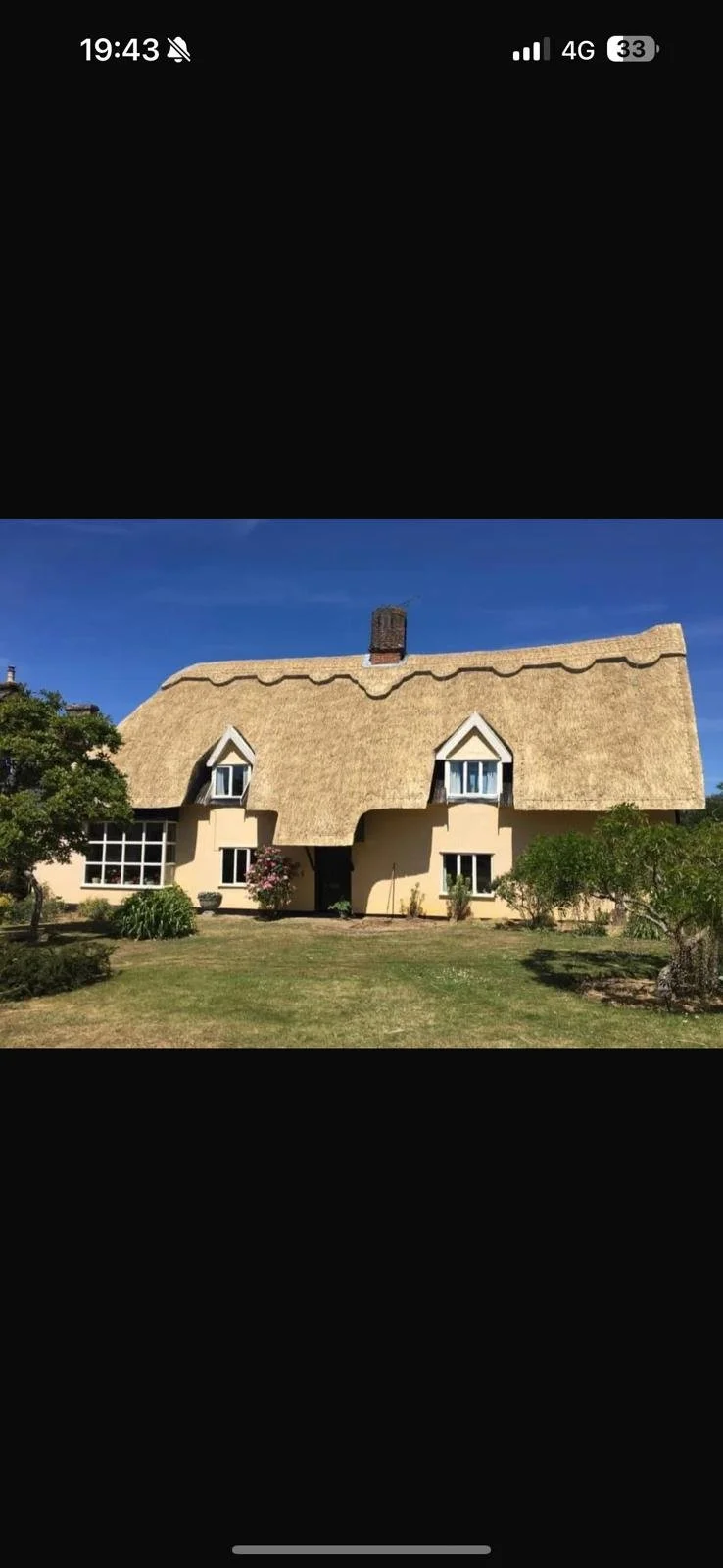 A house with a thatched roof, white walls, and four windows, surrounded by a lawn with trees and bushes, under a clear blue sky.