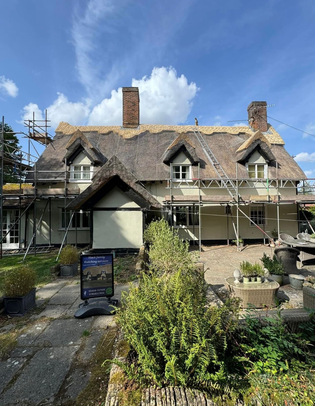 House under renovation with scaffolding, thatched roof, chimney, and a garden with plants and outdoor furniture.
