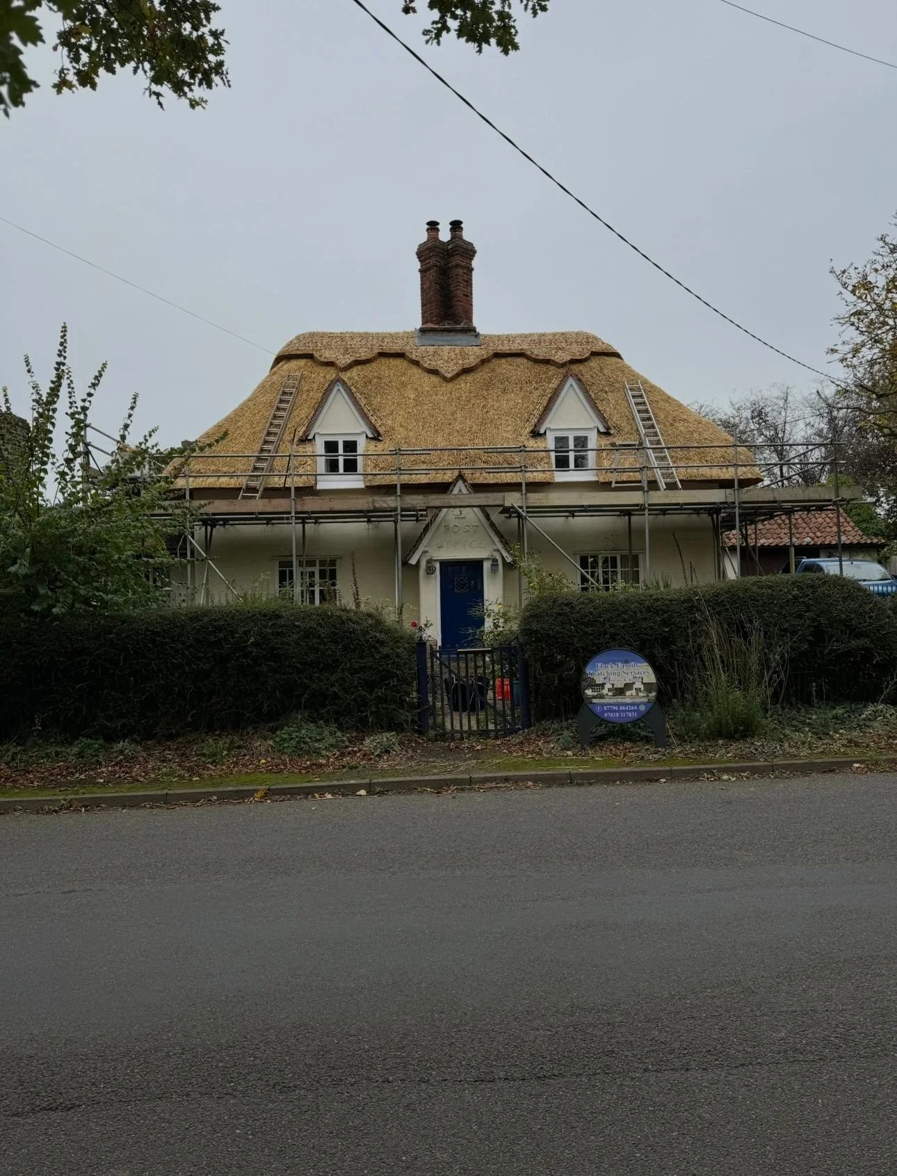 A house under construction with scaffolding, a thatched roof, and a blue front door, surrounded by bushes and trees.