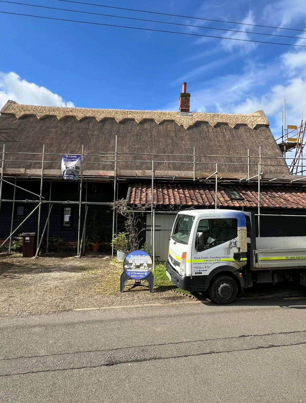 A thatched roof house under renovation with scaffolding and a service truck parked in front. The house has a bright blue sky background.