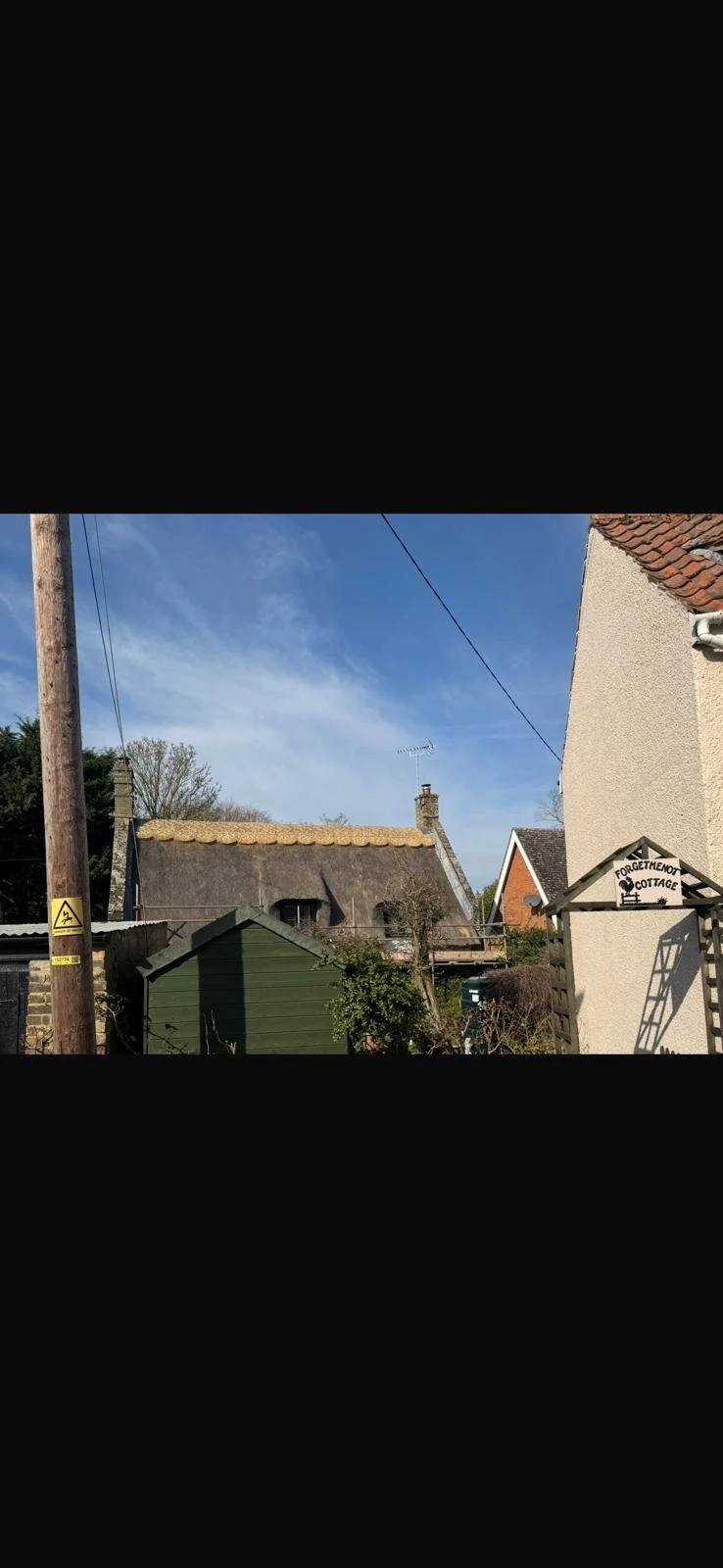 A cozy countryside scene featuring a thatched-roof cottage, a small green shed, and a sign reading "Forget-me-not Cottage" on an archway. There is a utility pole with wires, a blue sky with some clouds, trees, and neighboring houses.