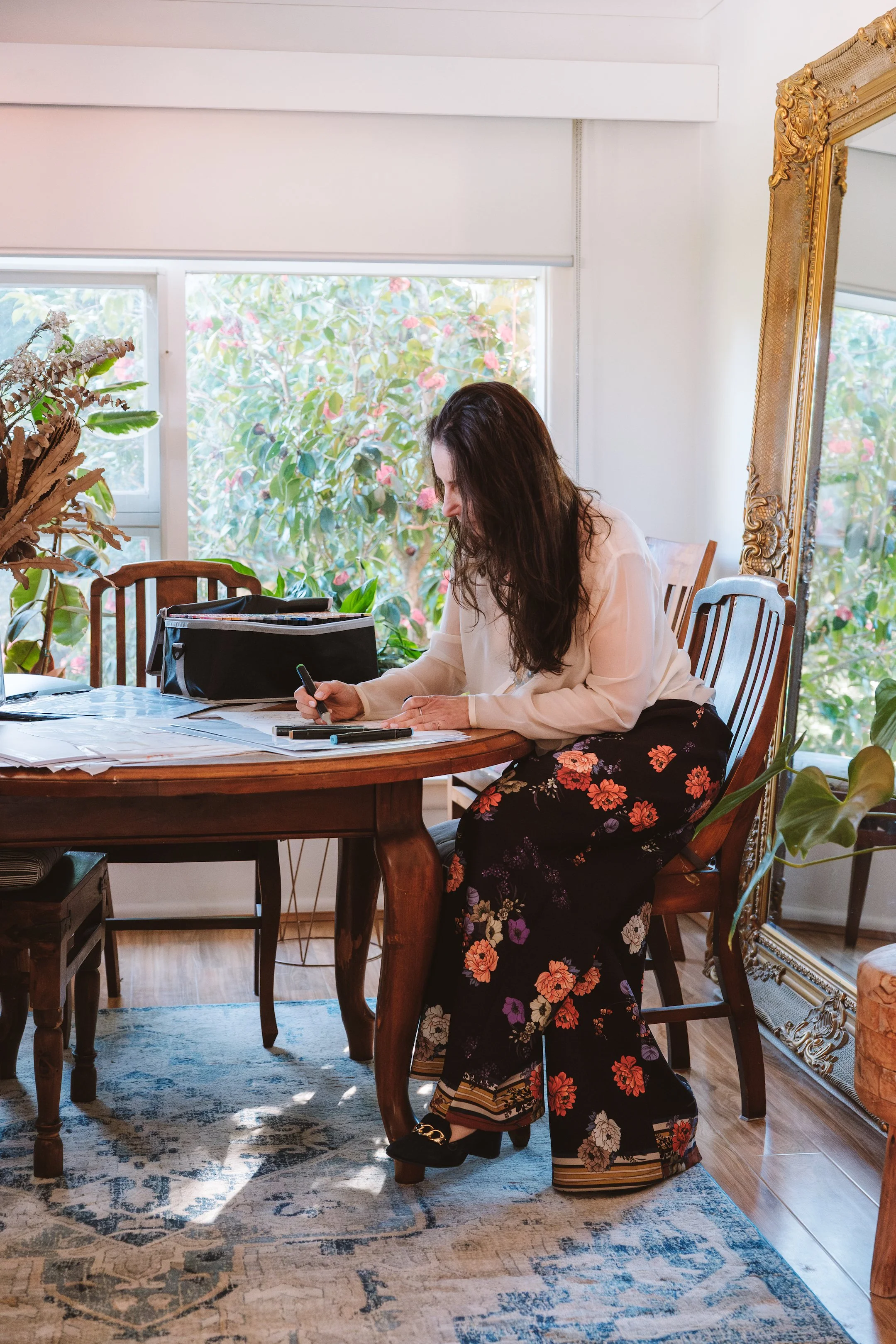 Woman with long brown hair sitting at a wooden table, writing on papers, in a room with plants, sunlight, and a large gold-framed mirror.