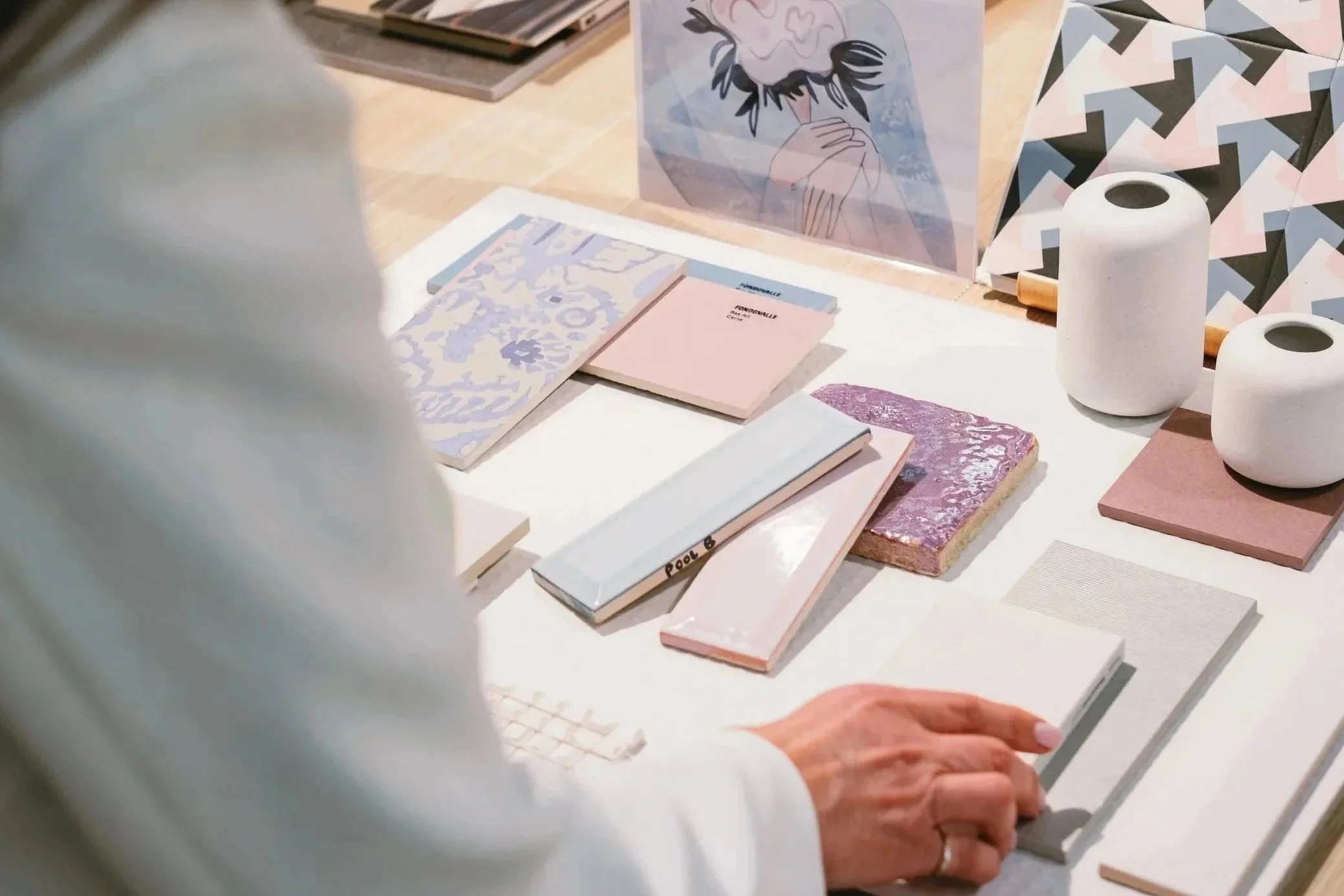 A person with light-colored clothing is selecting a rectangular tile from a display of various pastel-colored tiles and design samples, with patterned vases and decorative art in the background.