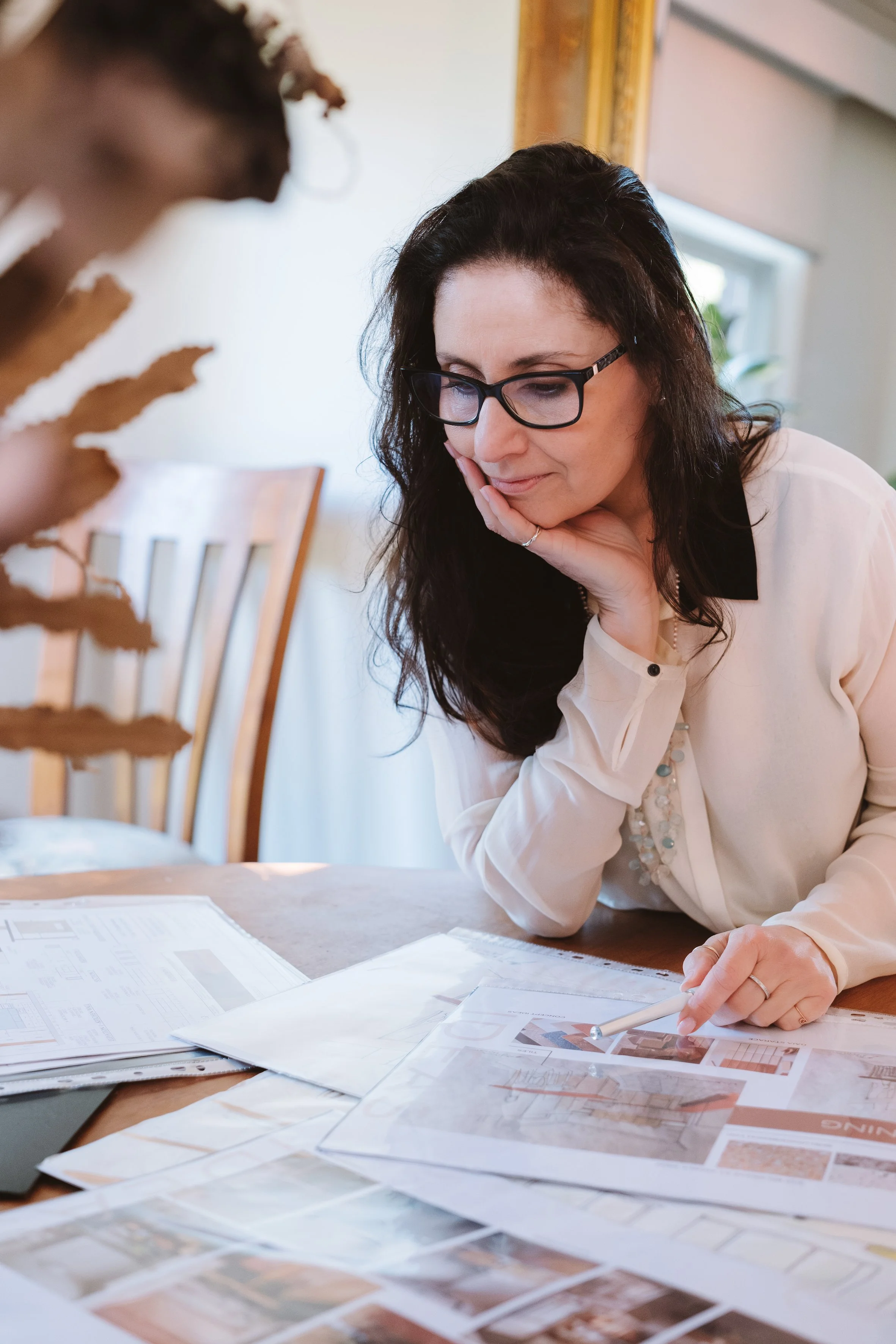 A woman with glasses, dark curly hair, wearing a light-colored blouse, is sitting at a table with various papers and color samples, looking at them thoughtfully.