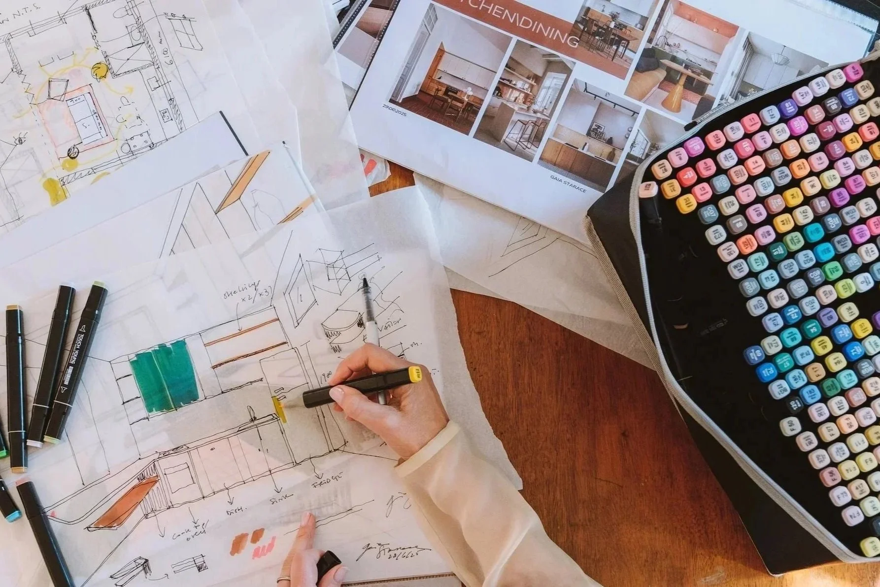 A person working on architectural sketches and interior design plans, with colored markers, design brochures, and a bag of colorful markers on a wooden table.