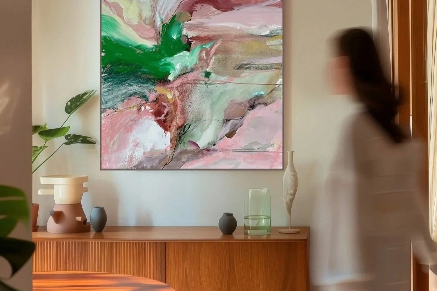 A modern interior with a large abstract painting featuring pink, green, and neutral tones on the wall. A wooden sideboard beneath the painting has decorative vases and a green glass container. A woman with long dark hair and a beige dress moves past on the right side, and a leafy plant with cut-out leaves is visible on the left.