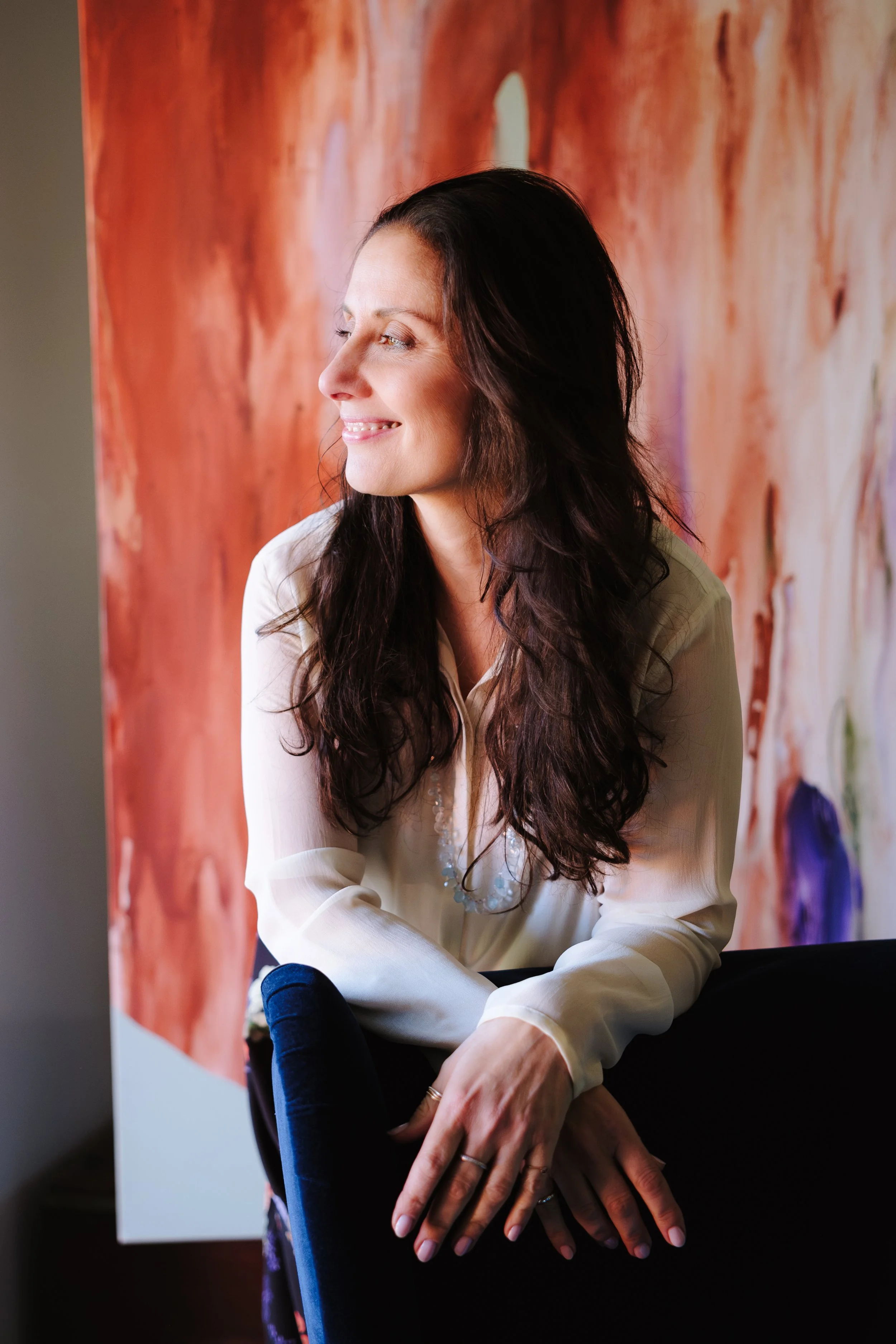 A woman with long dark hair, wearing a cream-colored blouse, sitting with her hands resting on her knee, looking out with a smile against a colorful abstract background.