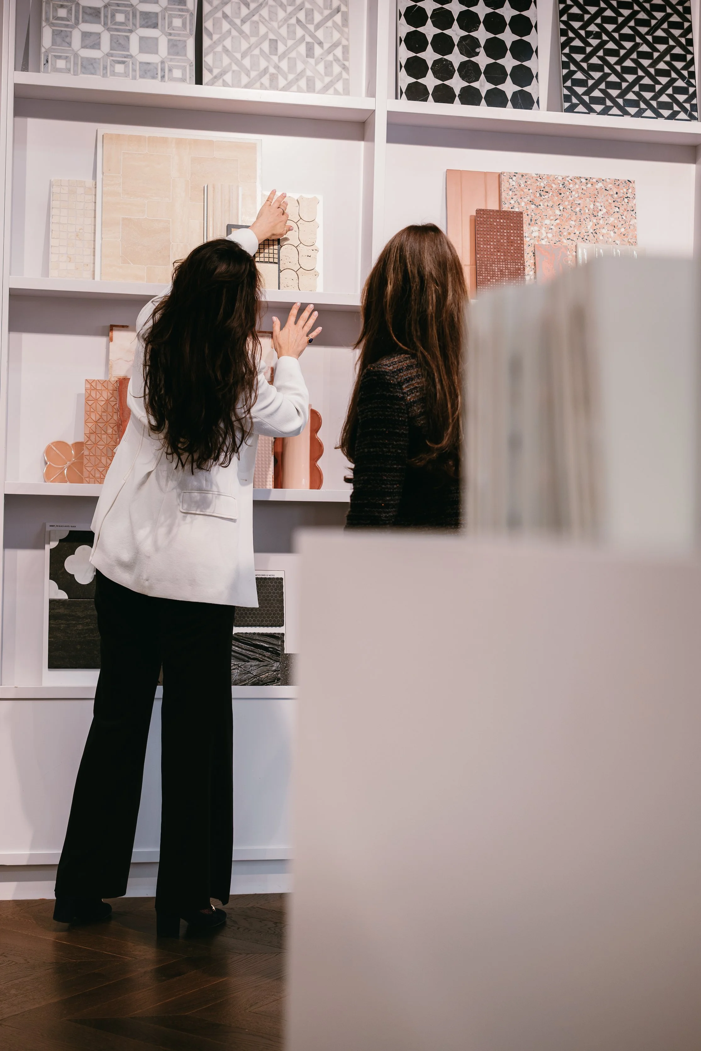 Two women looking at and discussing tile samples on display shelves in a store.