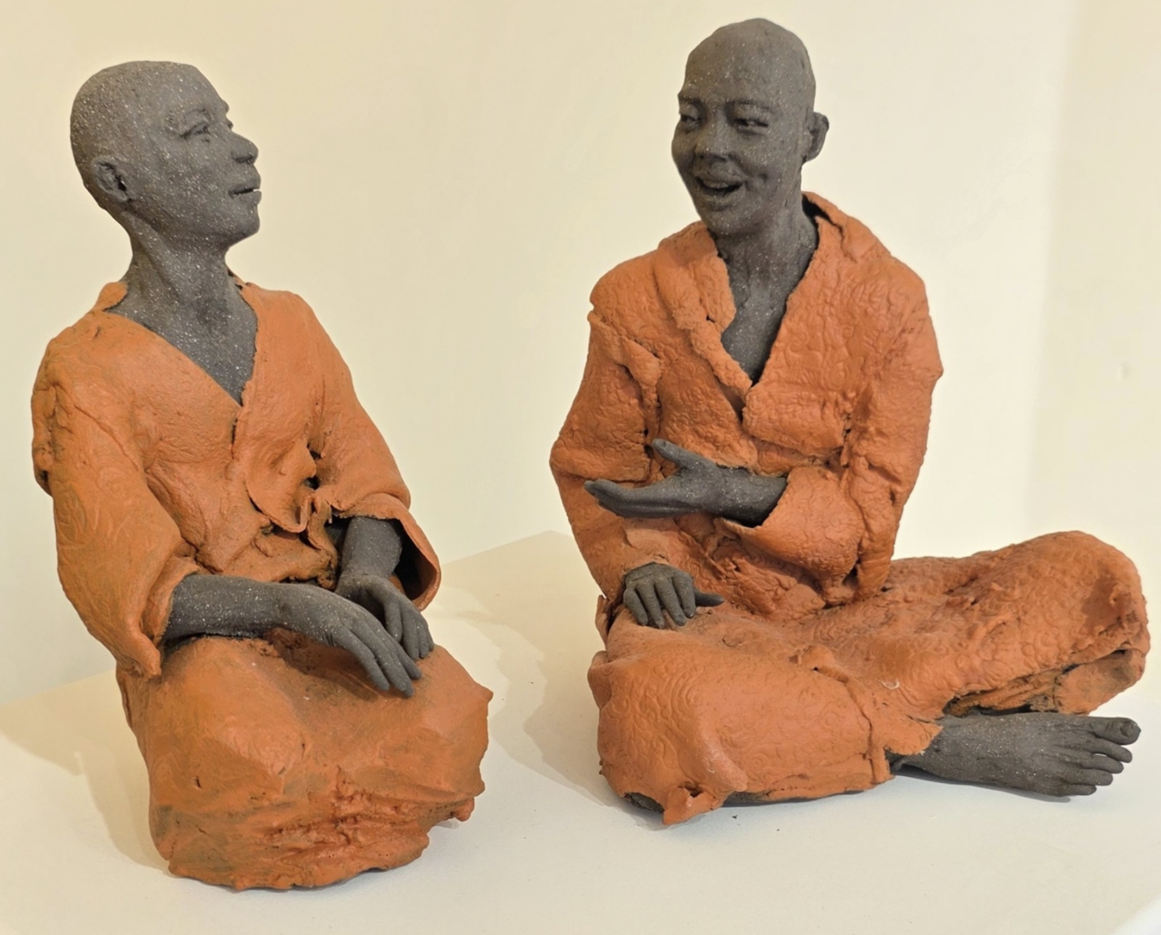 Deux sculptures en grès représentant deux personnes assises en conversation, portant des vêtements orange, avec un fond clair.