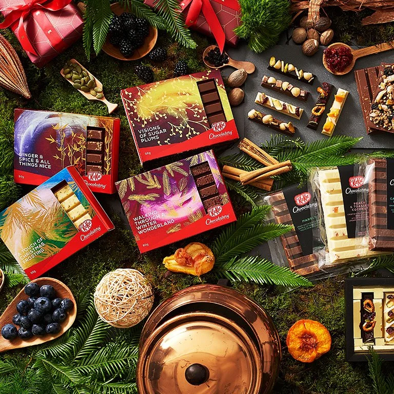 Assorted chocolates and candy bars with holiday decorations including pine branches, berries, cinnamon sticks, and dried fruits on a mossy green background.