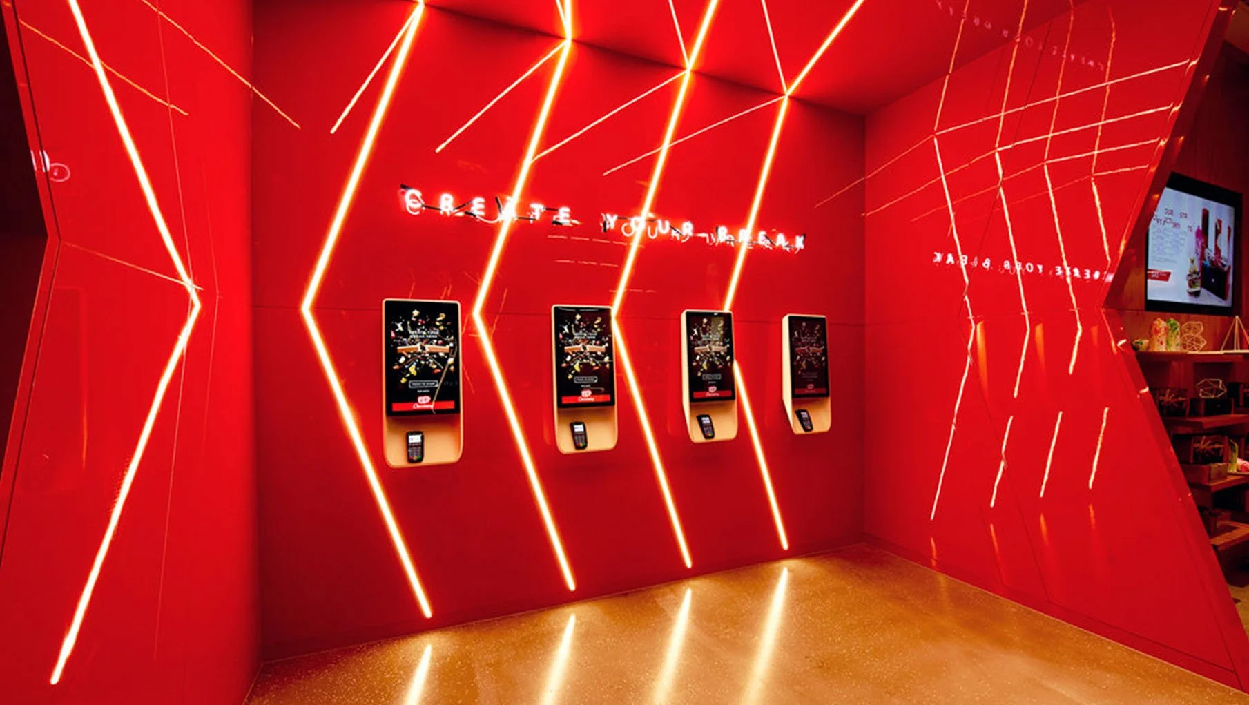 Four electronic kiosks with touch screens and card readers mounted on a red wall with diagonal red neon light strips. A partially visible red side wall is on the right. Red neon text above the kiosks reads 'Exercise Your Brain'.