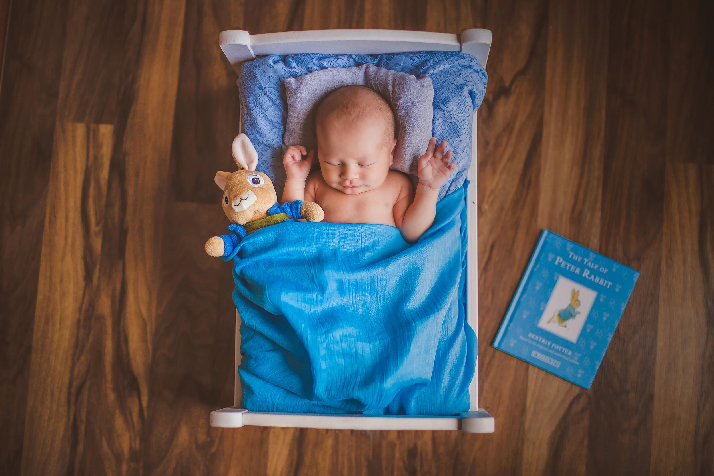An overhead view of a sleeping baby in a small bed with a blue blanket, a plush bunny toy, and a copy of 'The Tale of Peter Rabbit' book on a wooden floor.