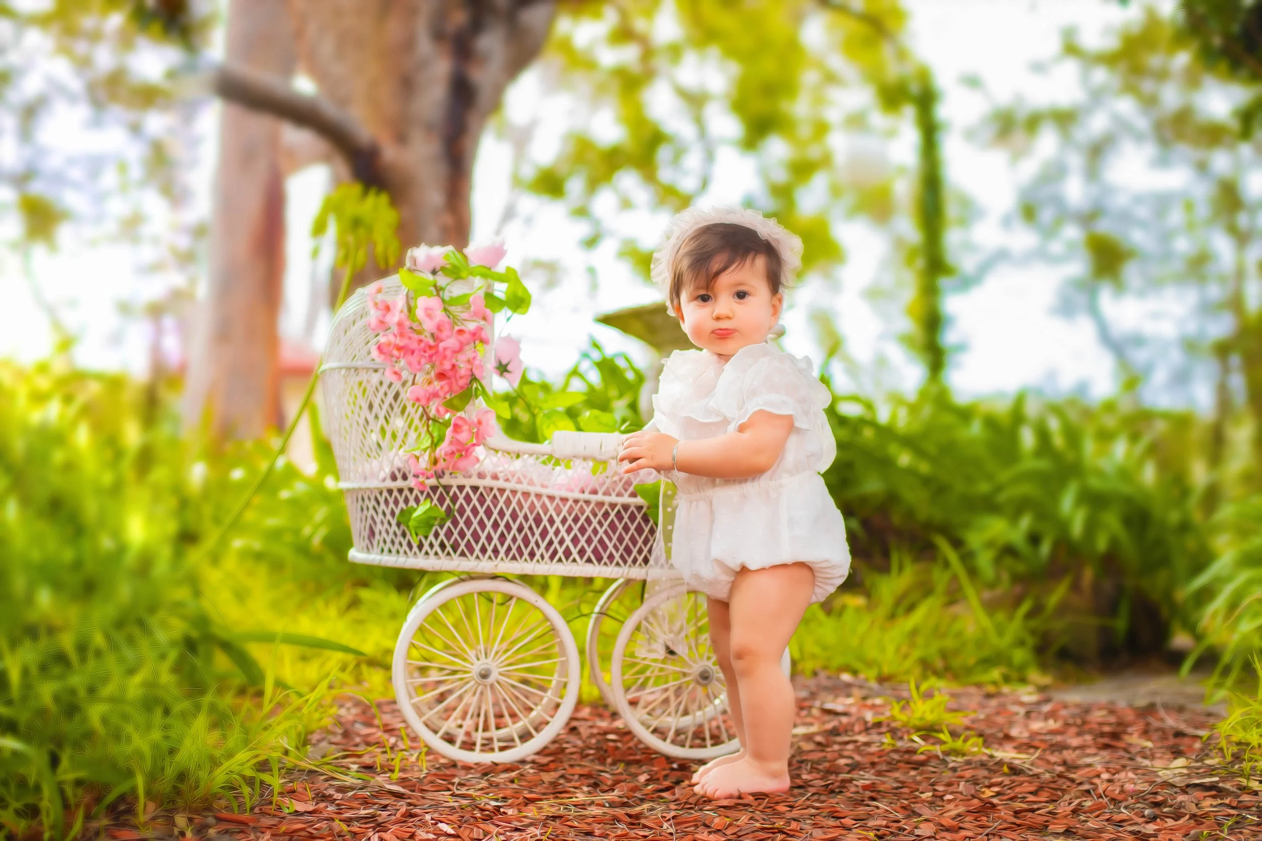 A young girl in a white dress standing next to a vintage white toy stroller with pink flowers, outdoors in a garden with trees and greenery.