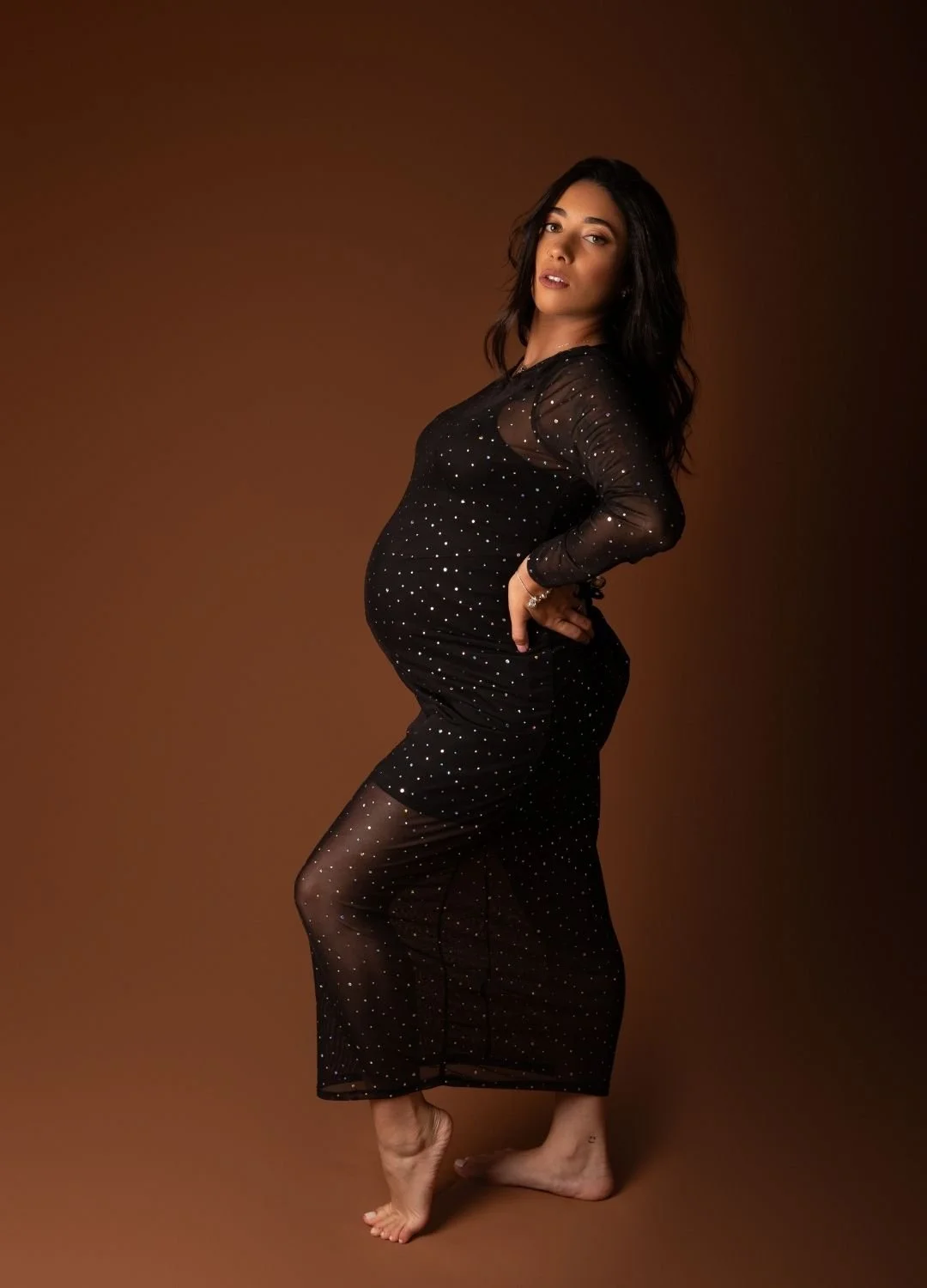 A pregnant woman with dark hair wearing a sheer black dress with small white polka dots, posing barefoot against a brown background.