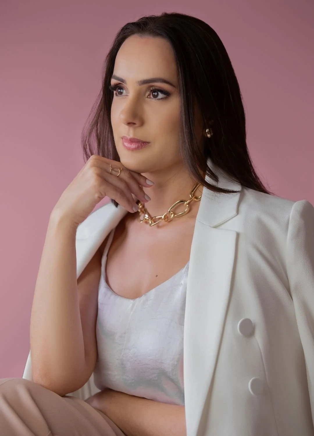 A woman with dark hair wearing a white blazer, a white top, a gold chain necklace, and earrings, sitting against a pink background, posing thoughtfully with her hand near her chin.