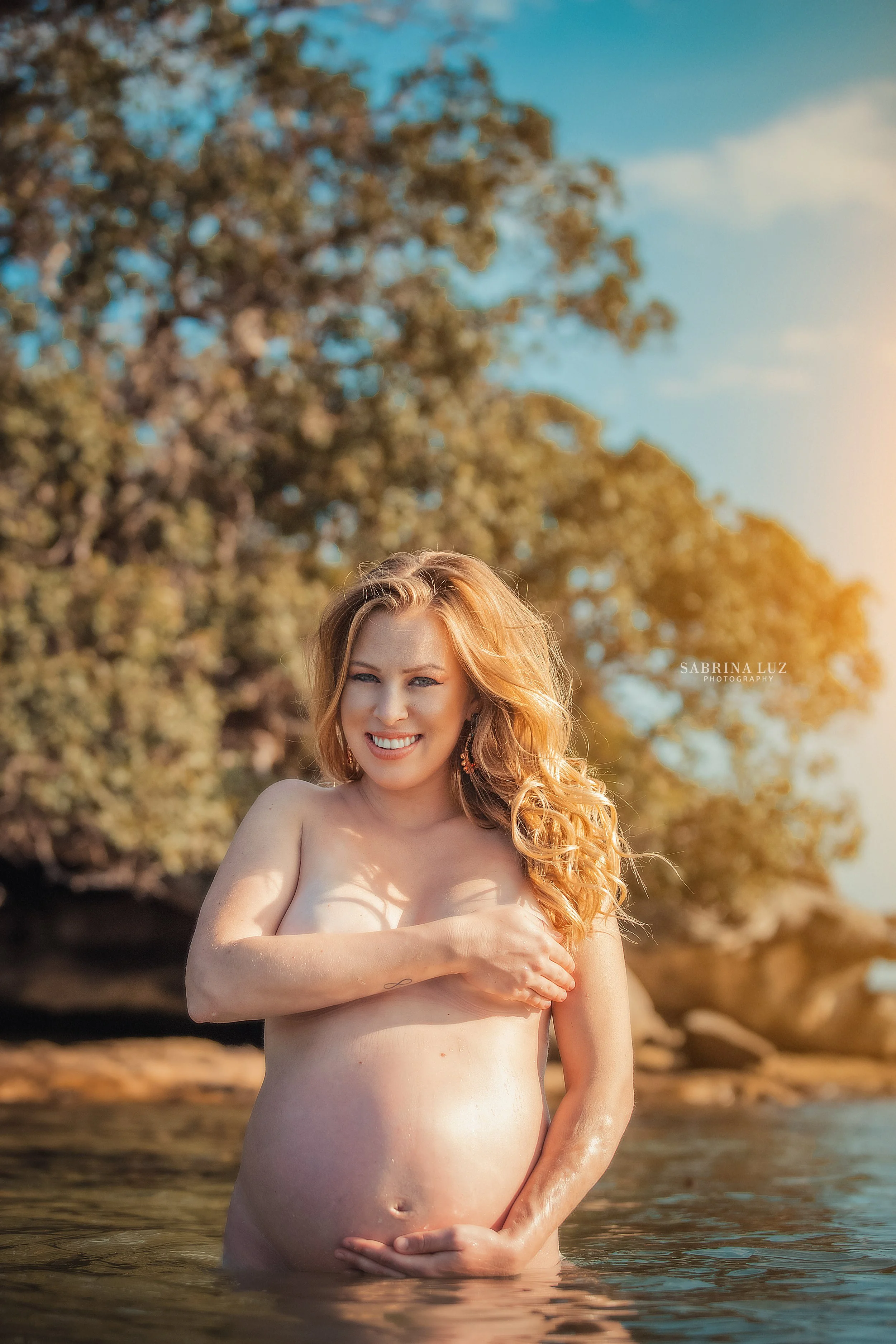 A pregnant woman standing in water outdoors, smiling and covering her chest with one hand, with a background of trees and a clear sky.