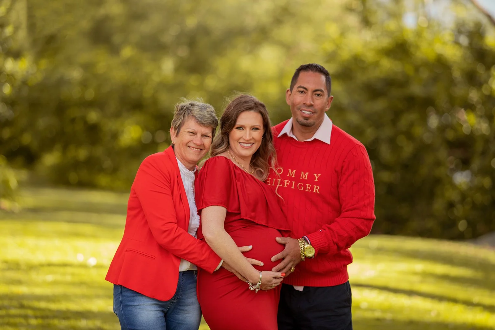 Three people, two women and one man, standing outdoors on a grassy area surrounded by trees, all dressed in red and smiling, with the woman in the middle being pregnant and cradling her belly.