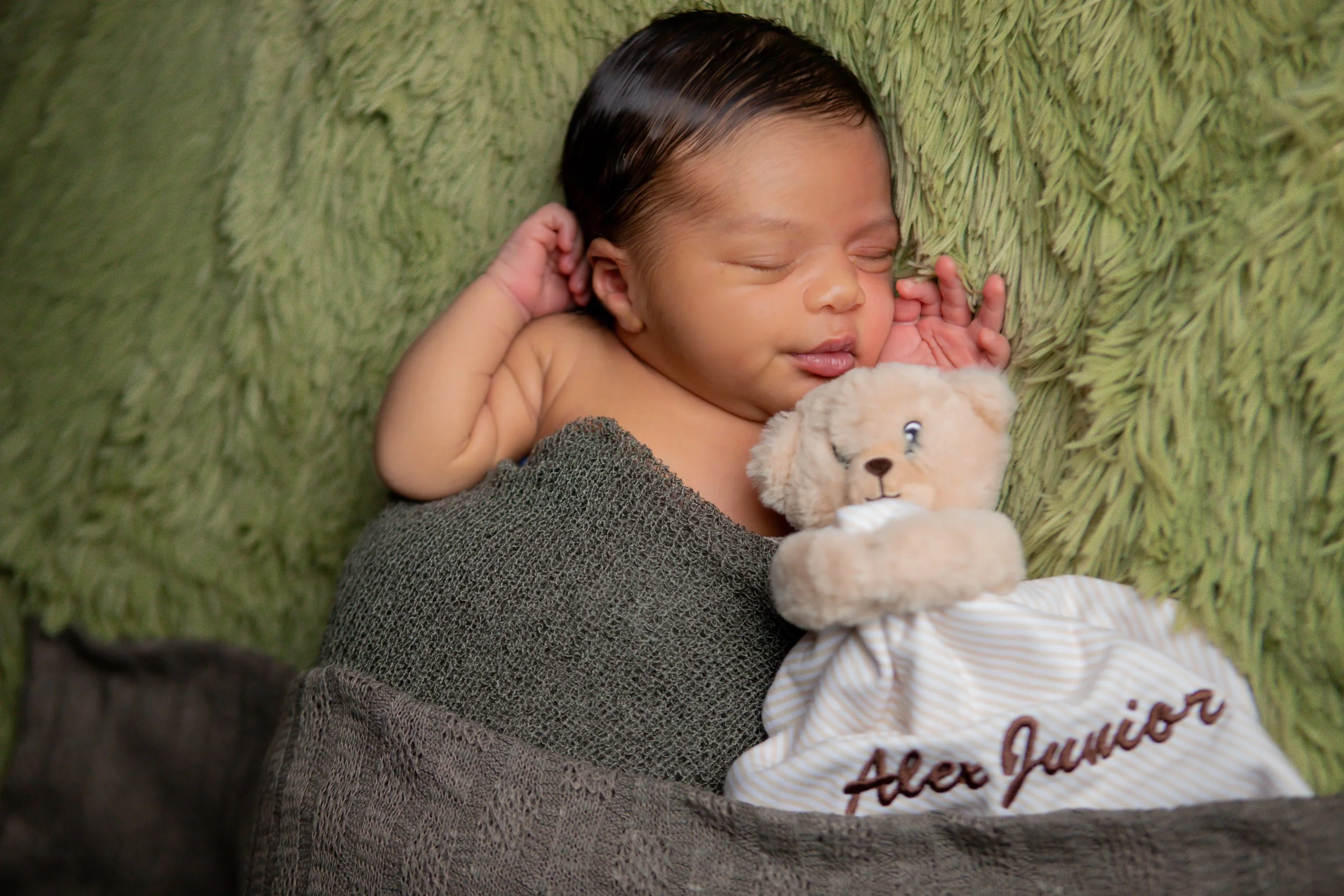 A sleeping baby with dark hair lies on a green fuzzy blanket, holding a beige teddy bear with a drawstring bag embroidered with 'Alder Junior'.