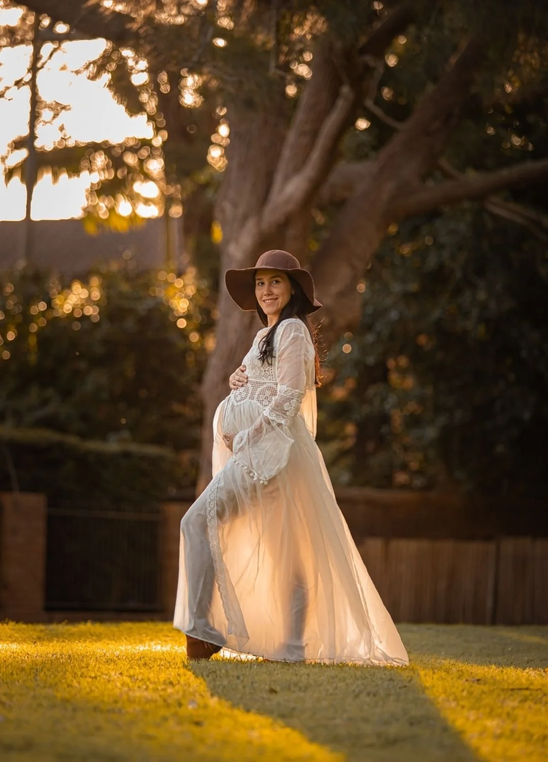 A pregnant woman in a long white dress and wide-brimmed hat standing outdoors at sunset, smiling and cradling her belly with her hands.